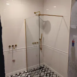 Nowoczesna kabina prysznicowa walk-in z białymi płytkami ściennymi, dekoracyjnymi panelami ściennymi, złotą armaturą i wzorzystą, czarno-białą podłogą, w trakcie montażu, widoczne niedokończone...