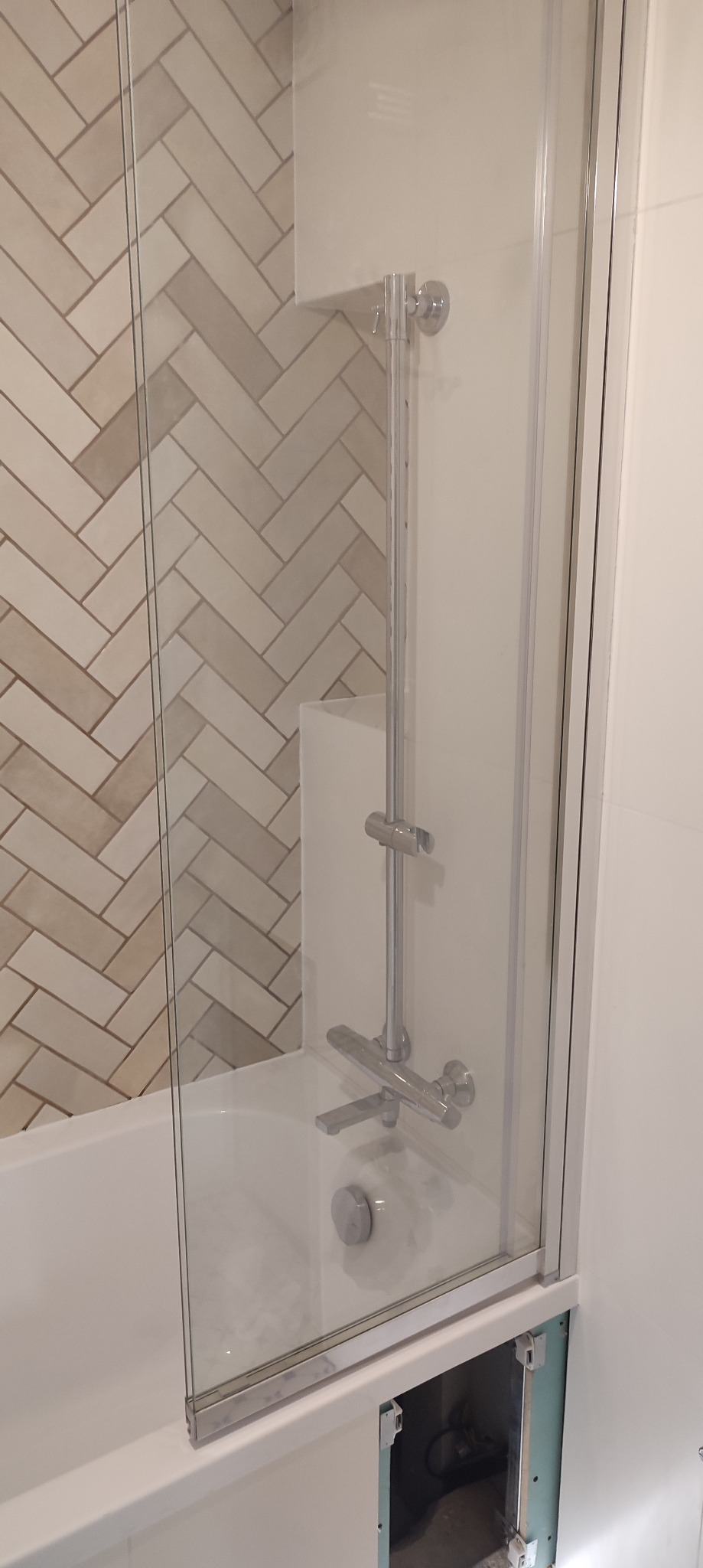 Szklany parawan nawannowy z chromowaną armaturą i płytkami ułożonymi w jodełkę, widoczny otwór rewizyjny pod wanną z widocznymi instalacjami.