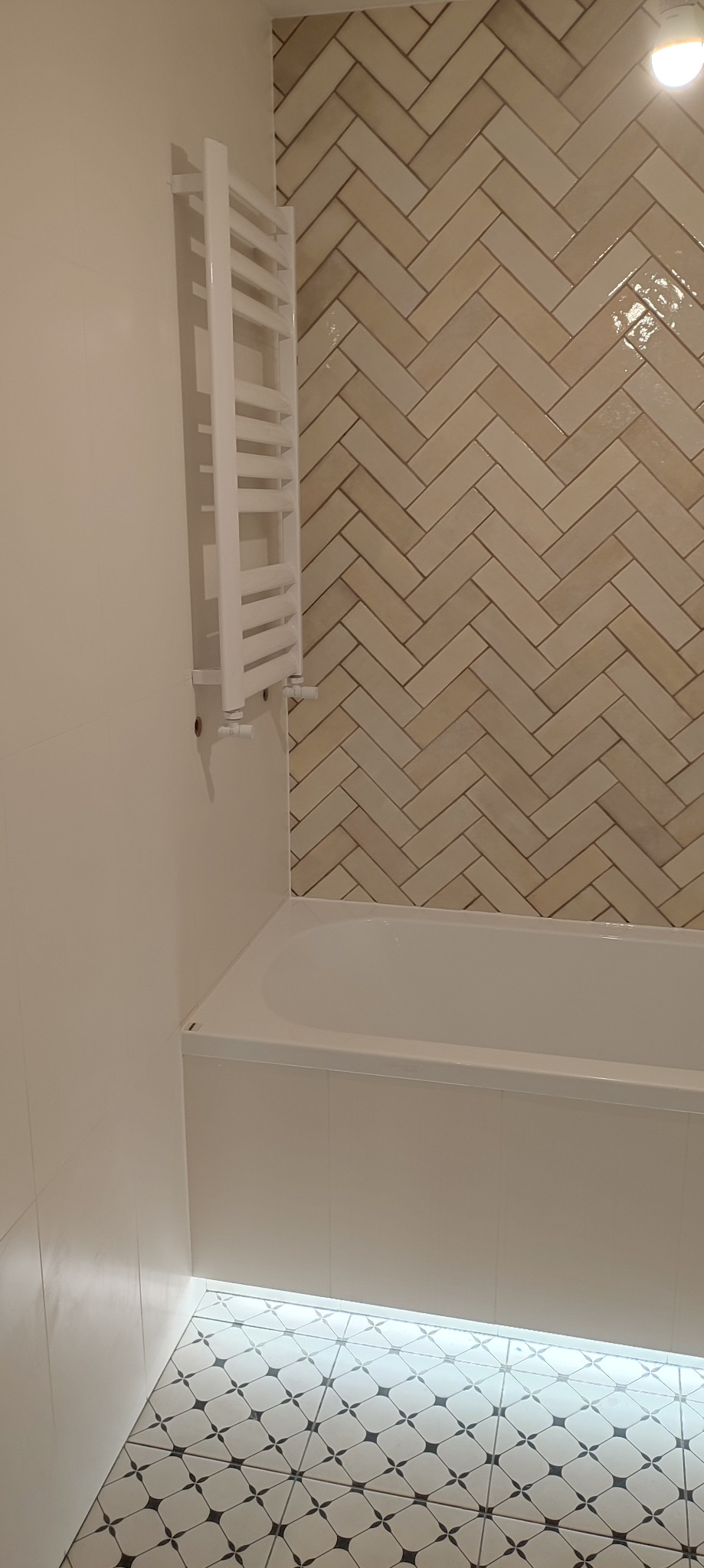 Wykończona łazienka w Raszynie: beżowa ściana z płytek ułożonych w jodełkę nad białą wanną, biały grzejnik drabinkowy i podłoga w czarno-białe wzory geometryczne z subtelnym oświetleniem LED przy...