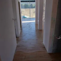 Wnętrze w trakcie prac wykończeniowych: ułożona podłoga z płytek imitujących drewno, surowe ściany z widocznymi przewodami elektrycznymi i kolumny w tle.