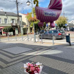 Bukiet różowych kwiatów leży na chodniku obok przejścia dla pieszych, a nad nim unosi się balon w kształcie wózka z napisem 'It's a Girl!', w tle ulica miejska z samochodami i budynkami.