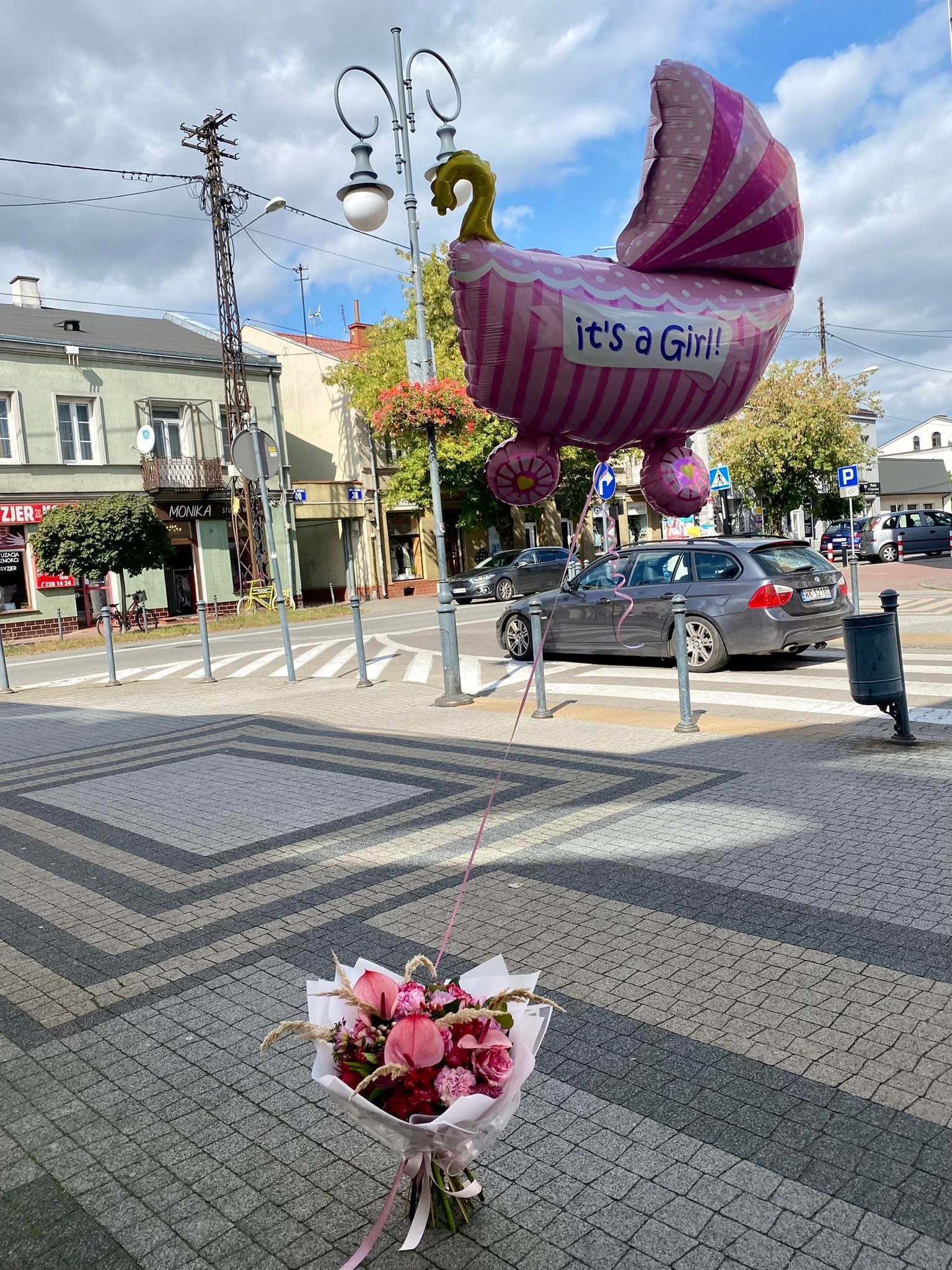 Bukiet różowych kwiatów leży na chodniku obok przejścia dla pieszych, a nad nim unosi się balon w kształcie wózka z napisem 'It's a Girl!', w tle ulica miejska z samochodami i budynkami.