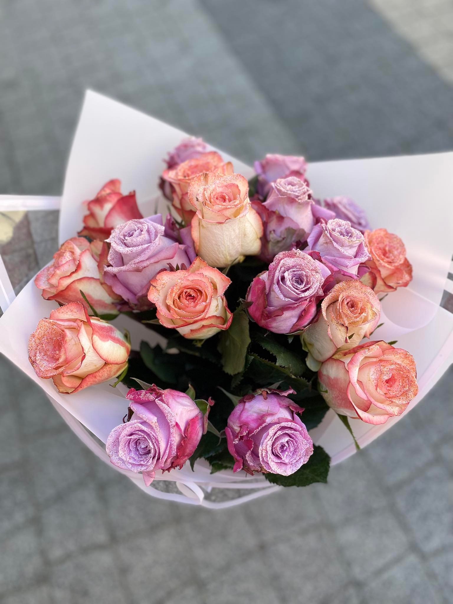 Bukiet mieszanych róż w odcieniach różu i fioletu, z delikatnym brokatem na płatkach, zapakowany w biały papier.