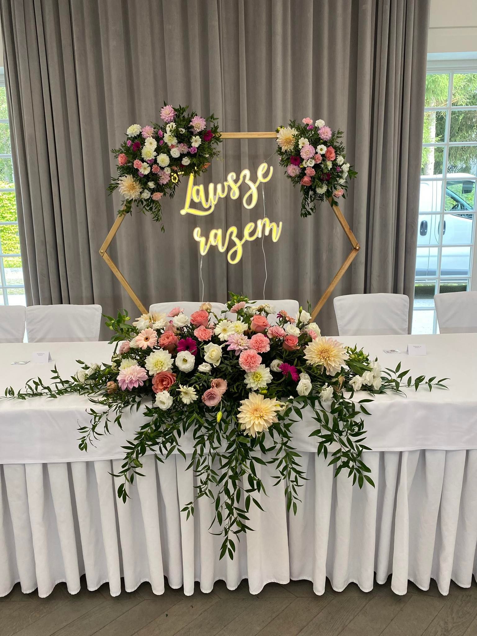 Dekoracja stołu weselnego z kompozycją kwiatową w odcieniach różu, bieli i kremu, z neonowym napisem 'Zawsze razem' w drewnianej ramie w kształcie heksagonu, na tle szarych zasłon i okna.