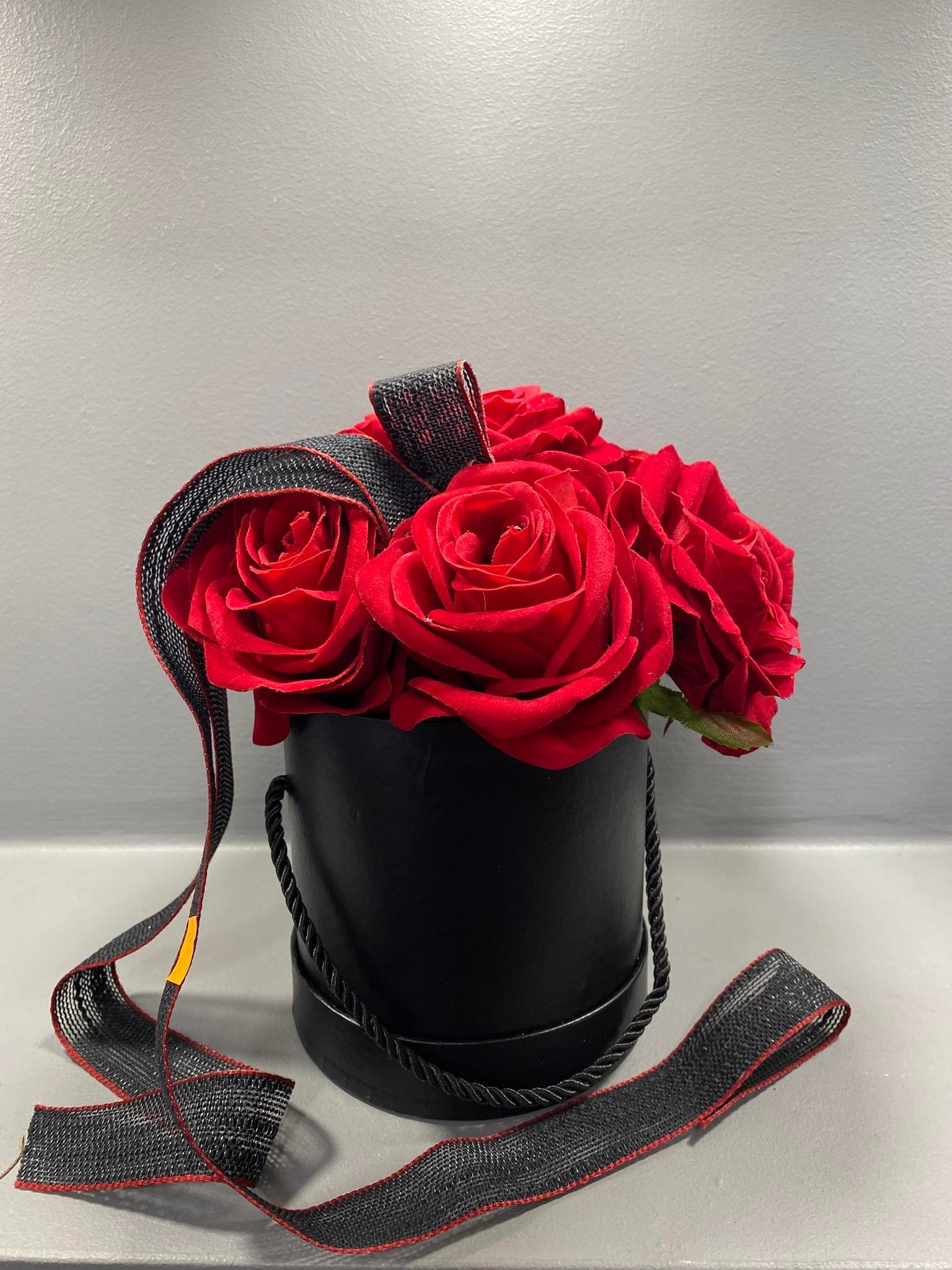Kompozycja z czerwonych róż w czarnym, okrągłym pudełku z czarną wstążką z czerwoną lamówką, na szarym tle.