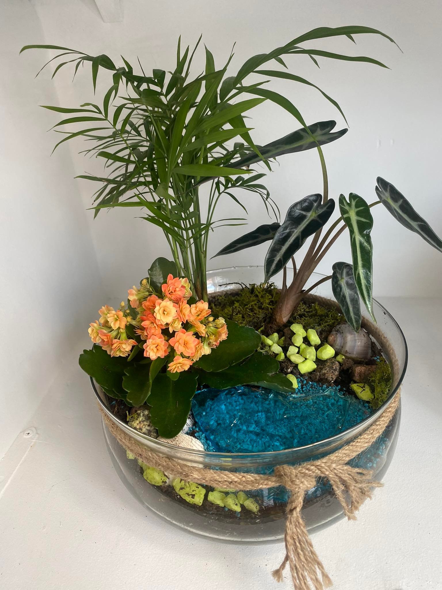 Szklane naczynie z kompozycją roślin, mchu, kamieni, muszli i turkusowego żwiru, ozdobione sznurkiem jutowym.