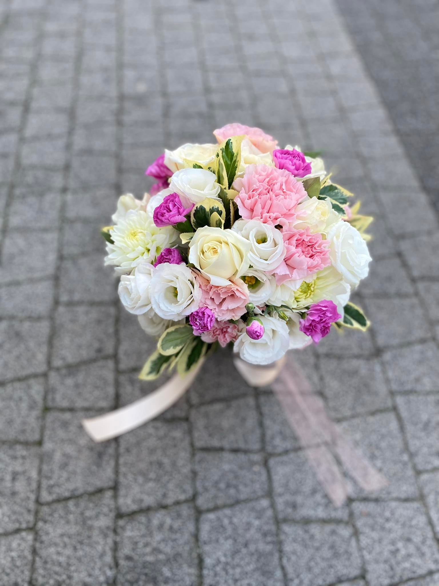 Bukiet z białych róż, różowych goździków i fioletowych eustom z dekoracyjną kremową wstążką, leżący na szarej kostce brukowej.