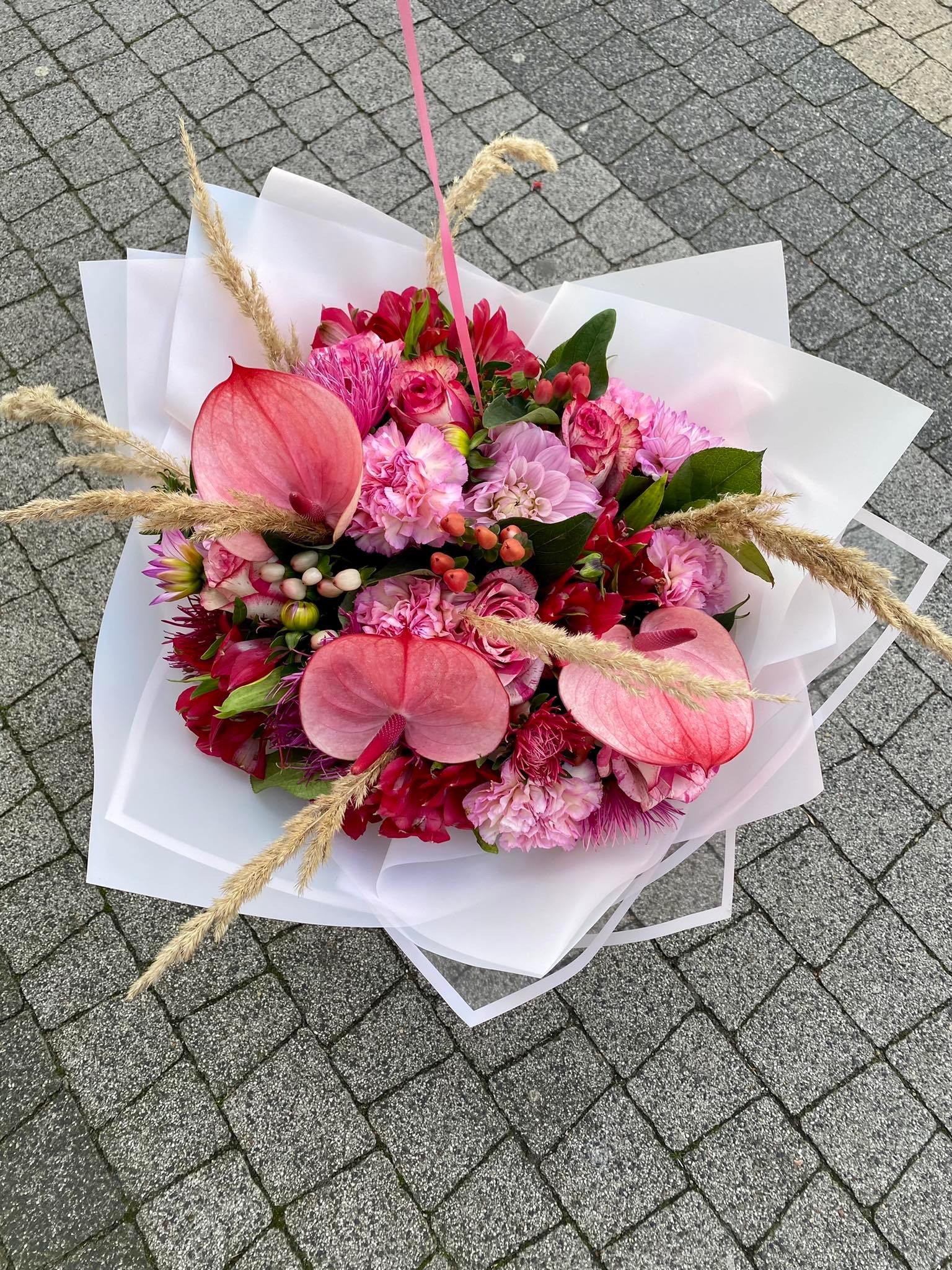 Widok z góry na bukiet różowych kwiatów anturium, róż i goździków, owinięty w biały papier i ozdobiony suchymi trawami, leżący na szarej kostce brukowej.