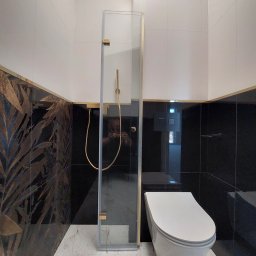 Jakub Waśkowski Remonty i Wykończenia - Nowoczesna łazienka z kabiną prysznicową ze złotymi detalami, czarnymi i białymi płytkami oraz marmurową podłogą. Widoczny element toalety podwieszanej.