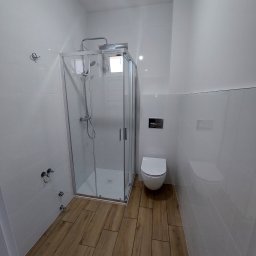 Jakub Waśkowski Remonty i Wykończenia - Nowoczesna łazienka z kabiną prysznicową, toaletą podwieszaną i drewnopodobną podłogą. Ściany wyłożone białymi, prostokątnymi płytkami.