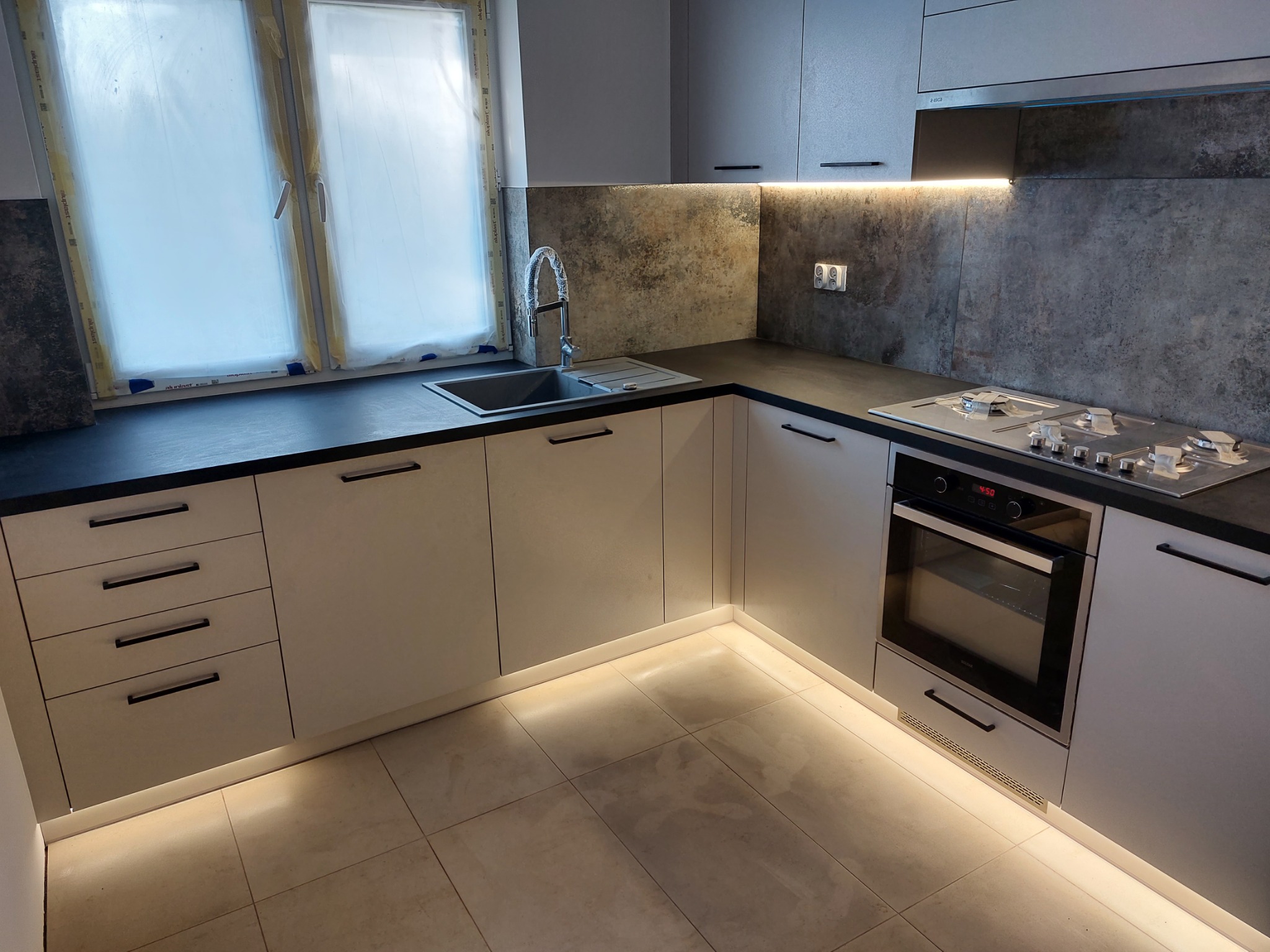 Nowoczesna kuchnia w odcieniach szarości z czarnym blatem, podświetleniem LED pod szafkami i nad blatem, oraz oknem zabezpieczonym folią ochronną.