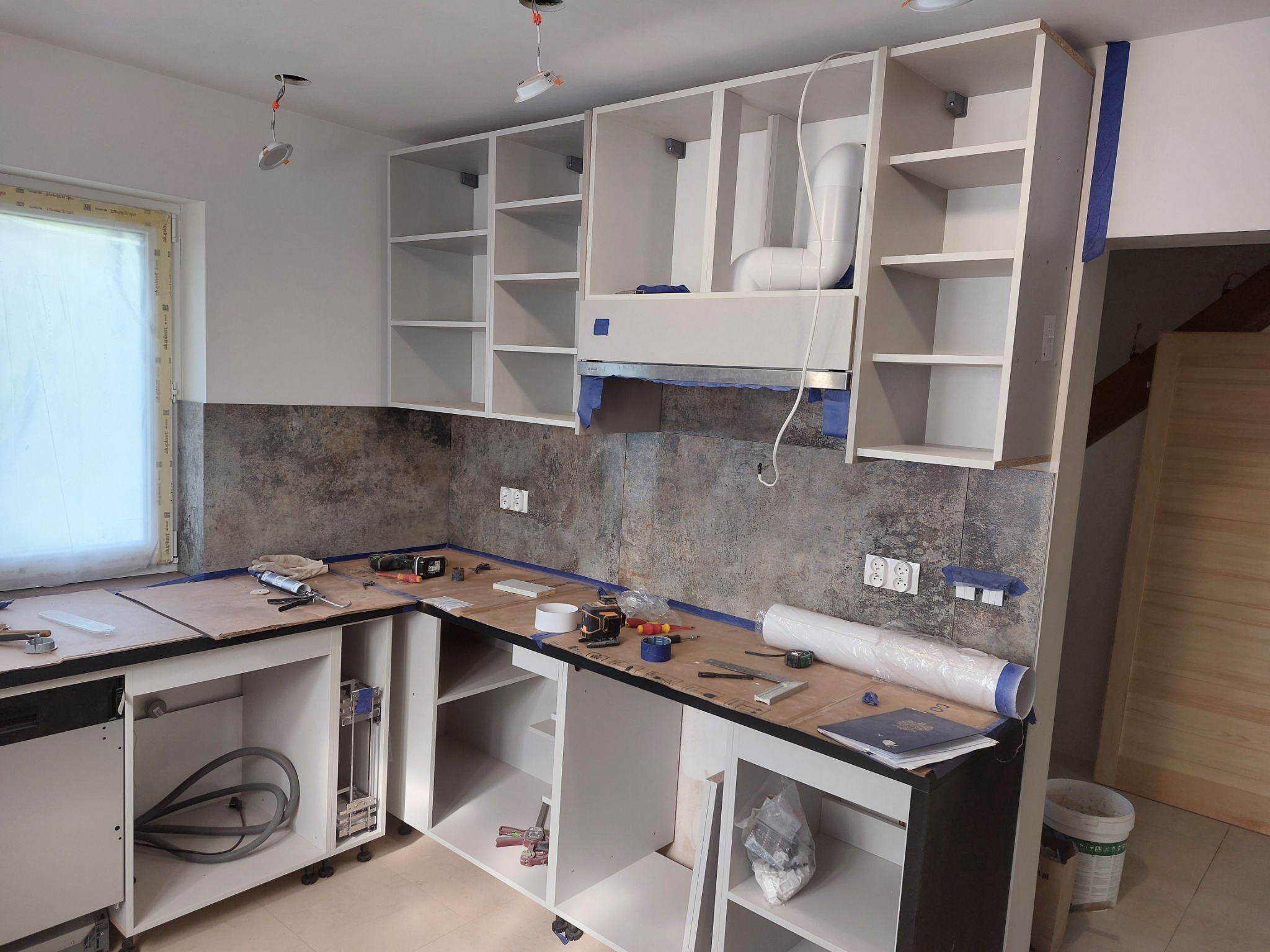 Montaż białych szafek kuchennych z szarym backsplashiem. Widoczne narzędzia na blacie, brakujące fronty szafek i niezakończone podłączenie oświetlenia sufitowego.