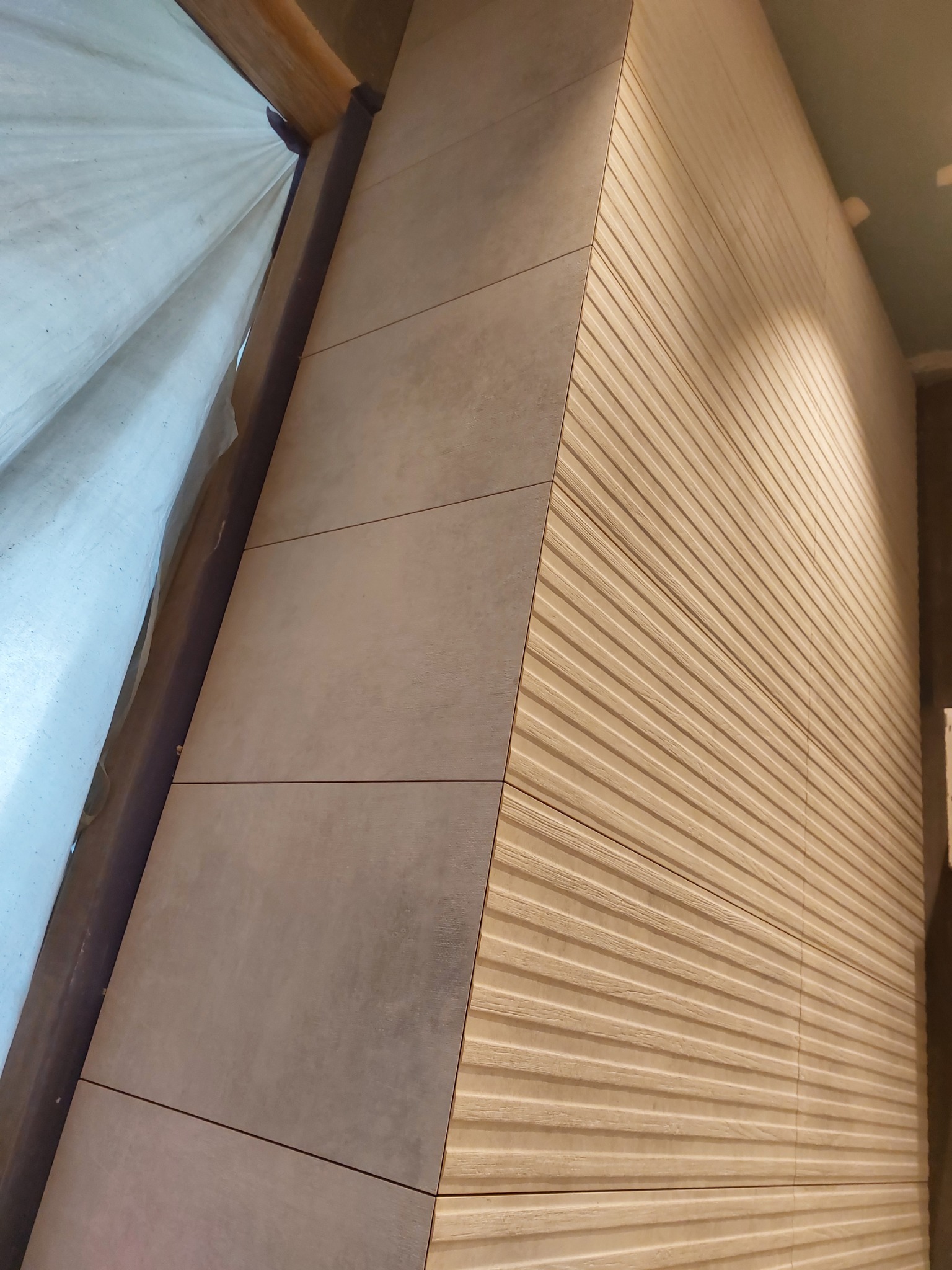 Narożnik ściany z fragmentem zakrytej folią budowlaną wnęki, wykończony płytkami ceramicznymi: z lewej gładkie, szare, z prawej beżowe z poziomymi żłobieniami imitującymi drewno.