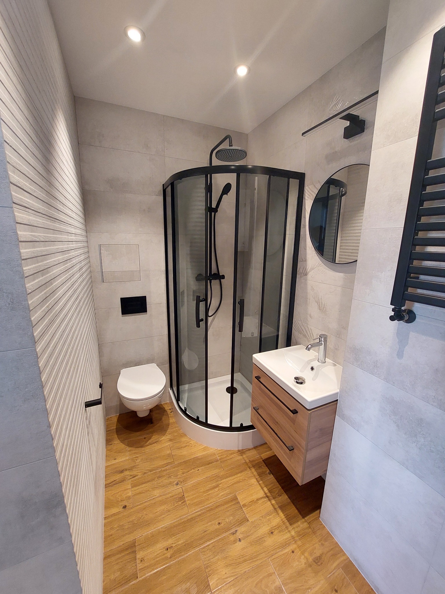 Nowoczesna łazienka z kabiną prysznicową typu walk-in z czarnymi profilami, umywalką nablatową na drewnianej szafce, podłogą imitującą drewno oraz ścianą z dekoracyjną fakturą.