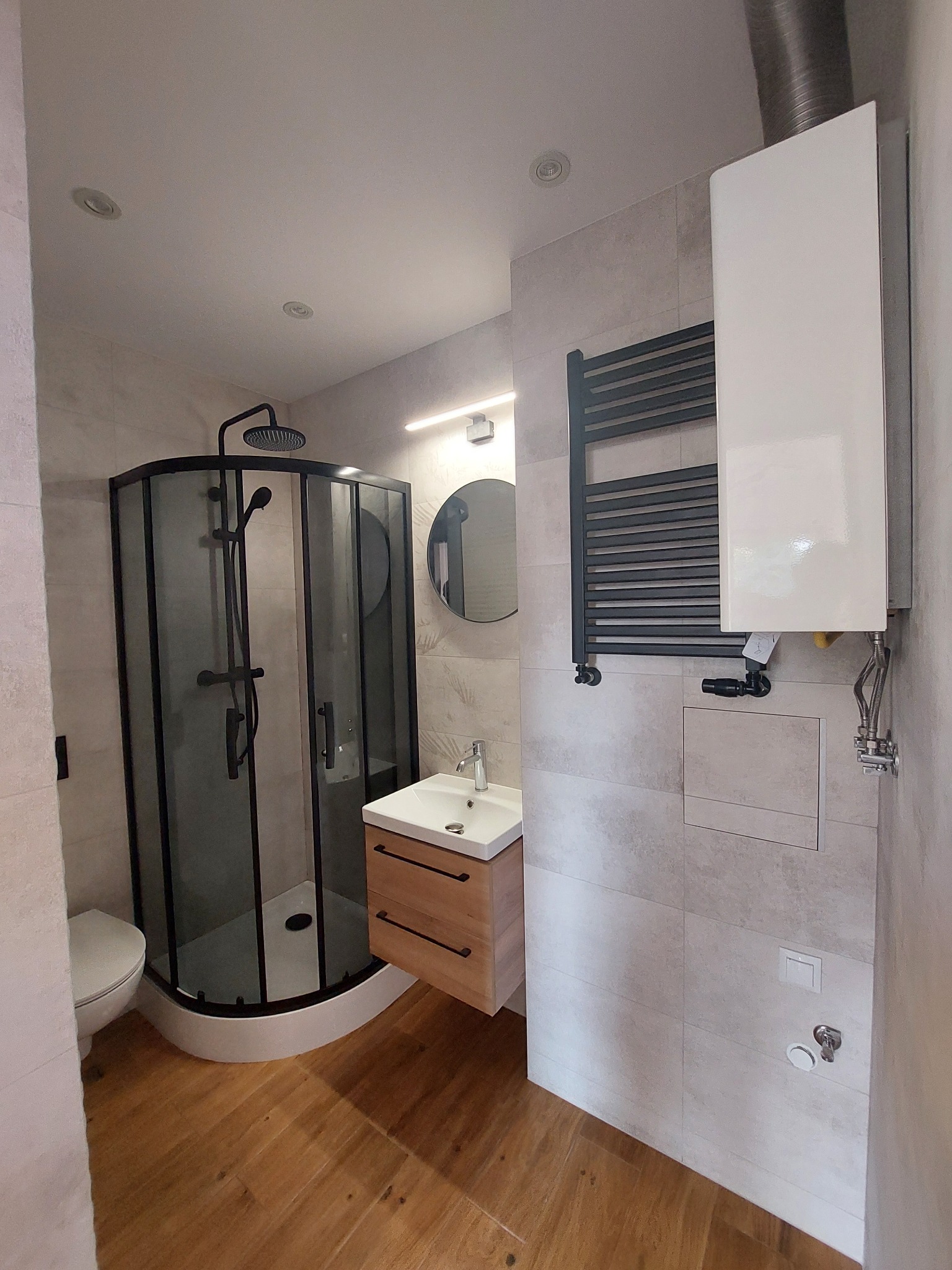 Nowoczesna łazienka z kabiną prysznicową typu walk-in z czarnymi ramami, drewnianą szafką pod umywalką z czarnymi uchwytami, grzejnikiem drabinkowym i białym bojlerem na ścianie z jasnych płytek.