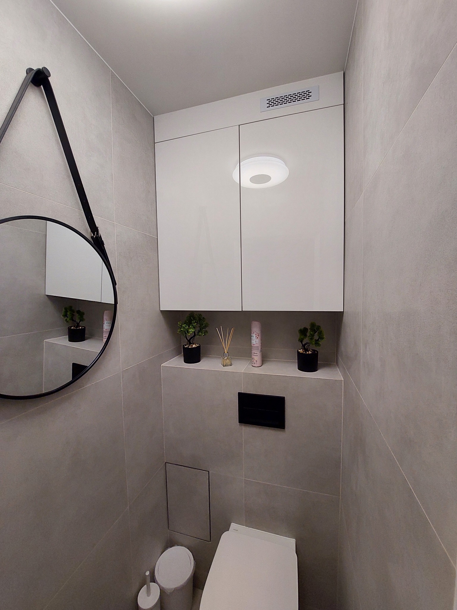Nowoczesna, minimalistyczna toaleta wykończona w szarych płytkach z wiszącą szafką, okrągłym lustrem na pasku i dekoracyjnymi roślinami w doniczkach.
