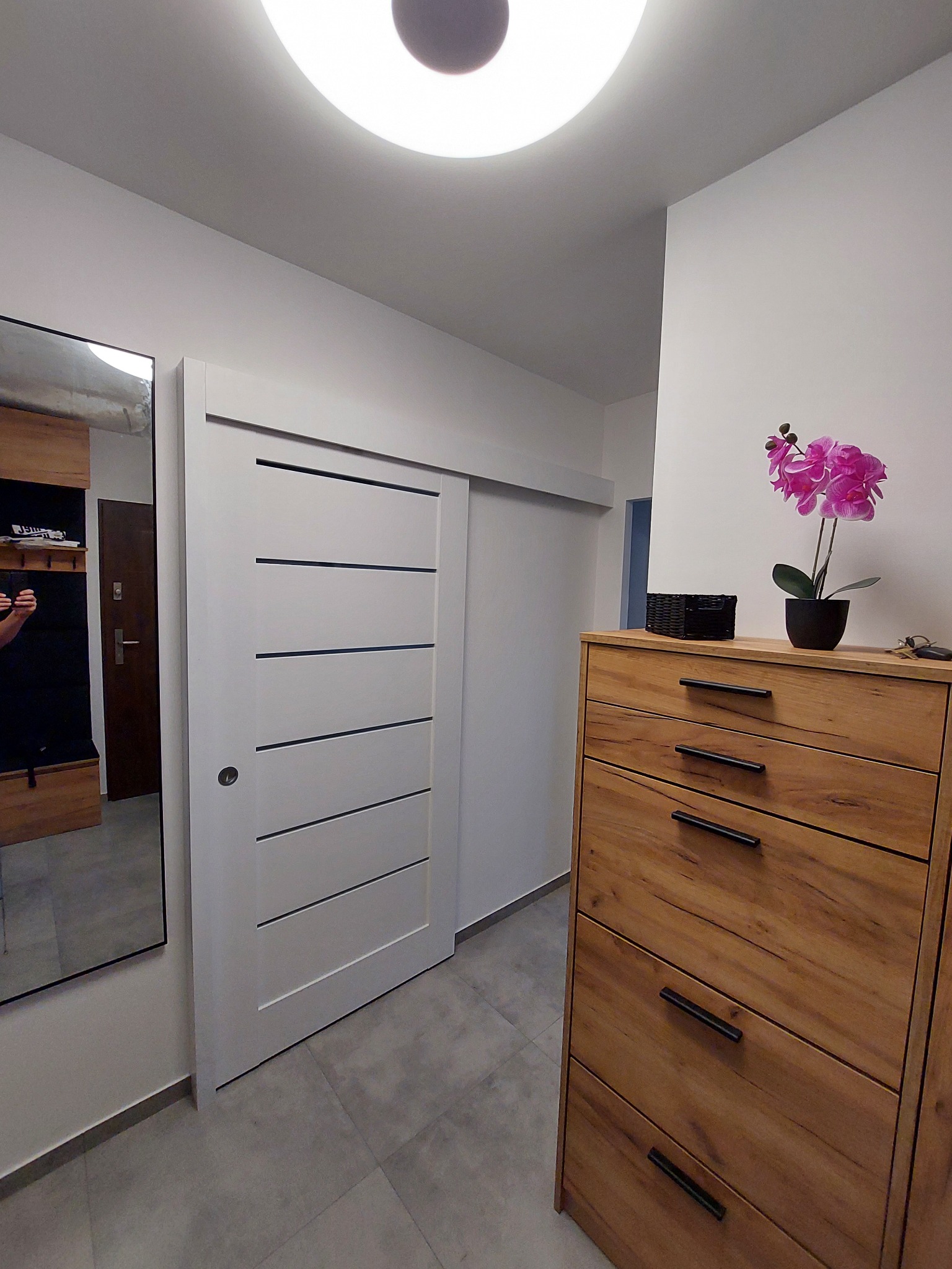 Nowoczesny przedpokój z przesuwnymi drzwiami w kolorze białym z poziomymi frezowaniami, drewniana komoda z czarnymi uchwytami, szara podłoga z płytek i odbicie osoby robiącej zdjęcie w lustrze.