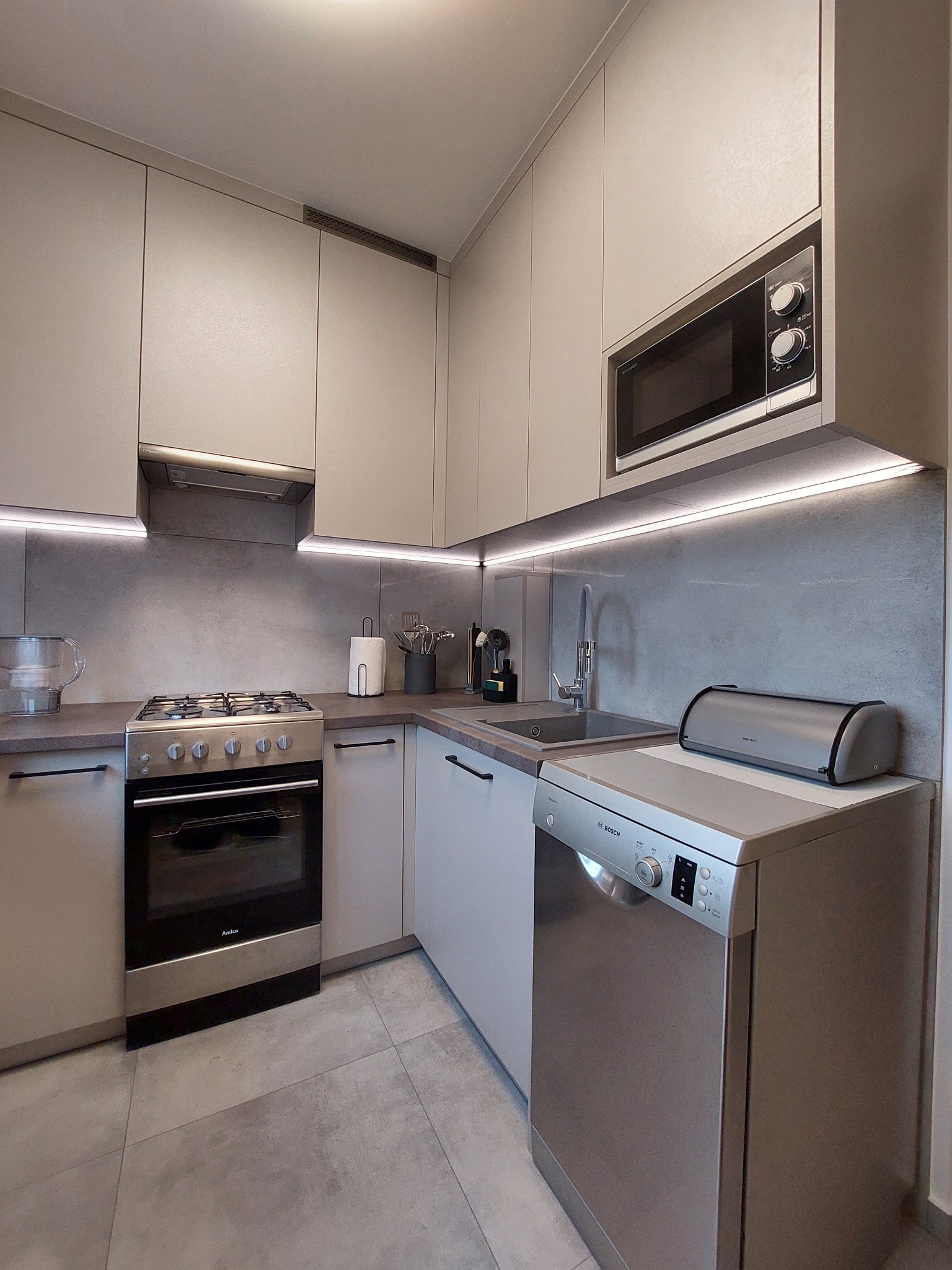 Nowoczesna kuchnia w odcieniach szarości z oświetleniem LED podszafkowym, sprzęt AGD marki Amica i Bosch, oraz szarym kamiennym blatem i płytkami podłogowymi.