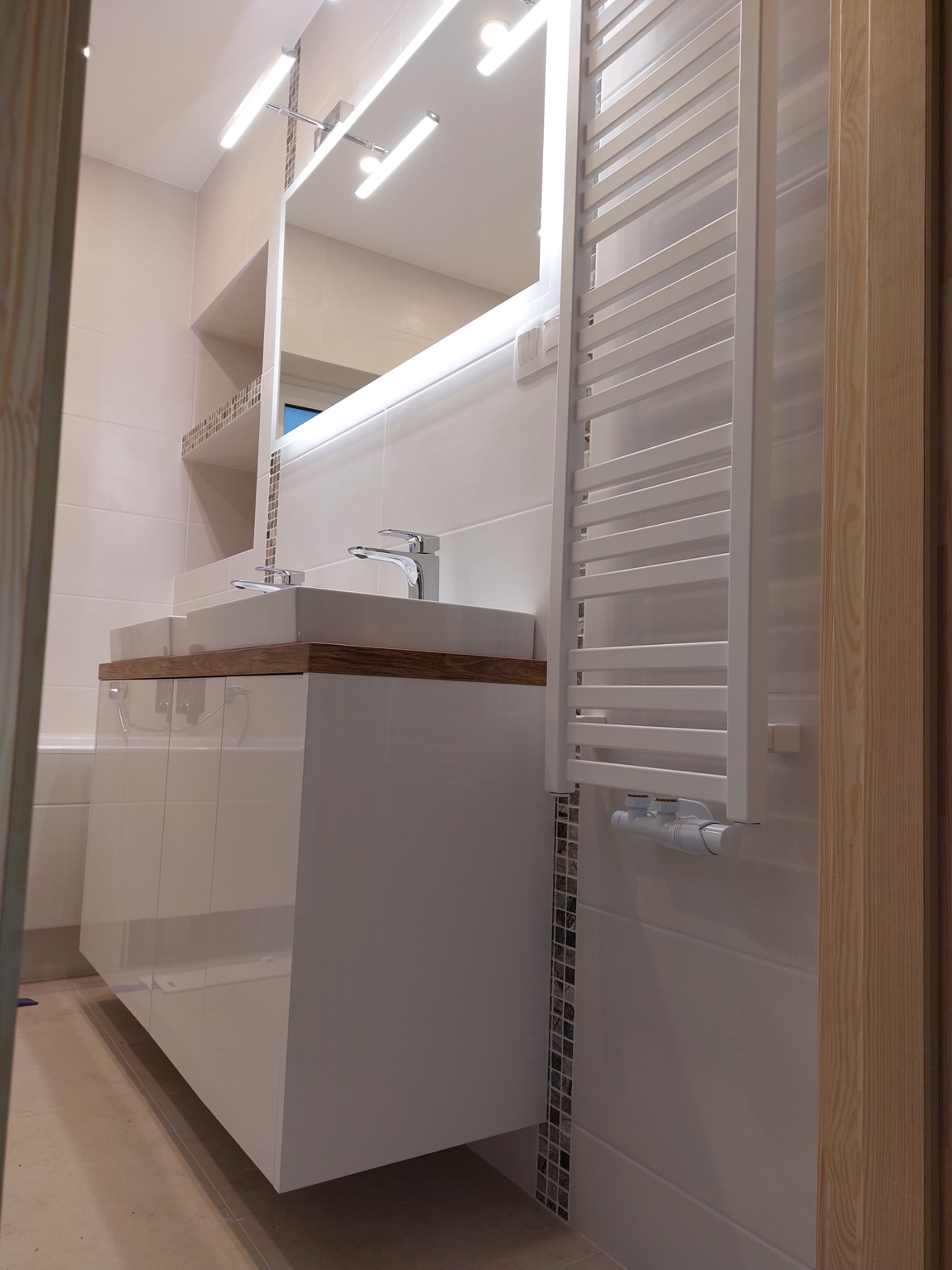 Nowoczesna łazienka z podwójną umywalką nablatową, szafką z białym połyskiem, grzejnikiem drabinkowym i oświetleniem LED nad lustrem.