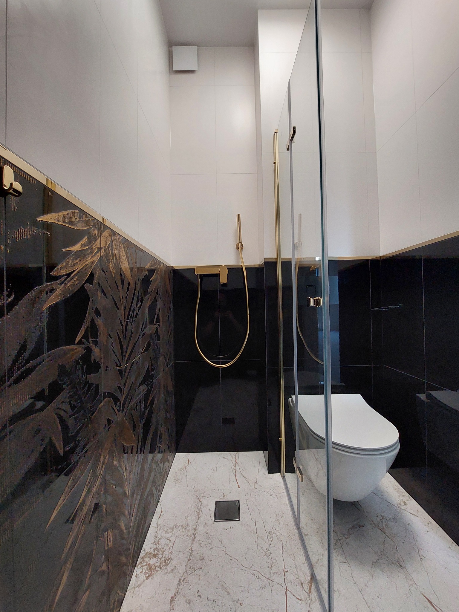 Nowoczesna łazienka z czarno-białymi płytkami, złotymi akcentami i kabiną prysznicową ze szkła. Na podłodze marmurowe płytki. Widoczny fragment toalety.