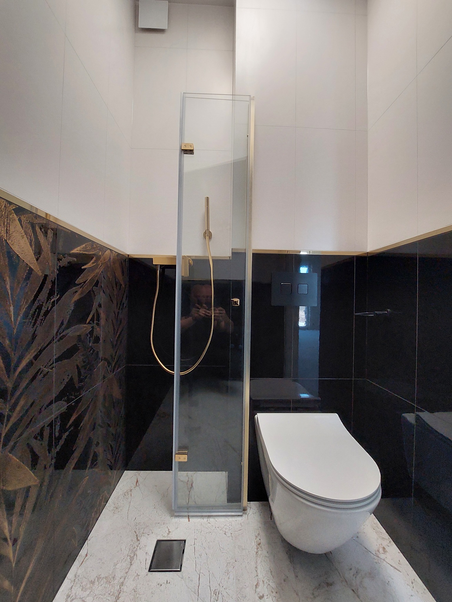 Nowoczesna łazienka z kabiną prysznicową ze złotymi detalami, czarnymi i białymi płytkami oraz marmurową podłogą. Widoczny element toalety podwieszanej.