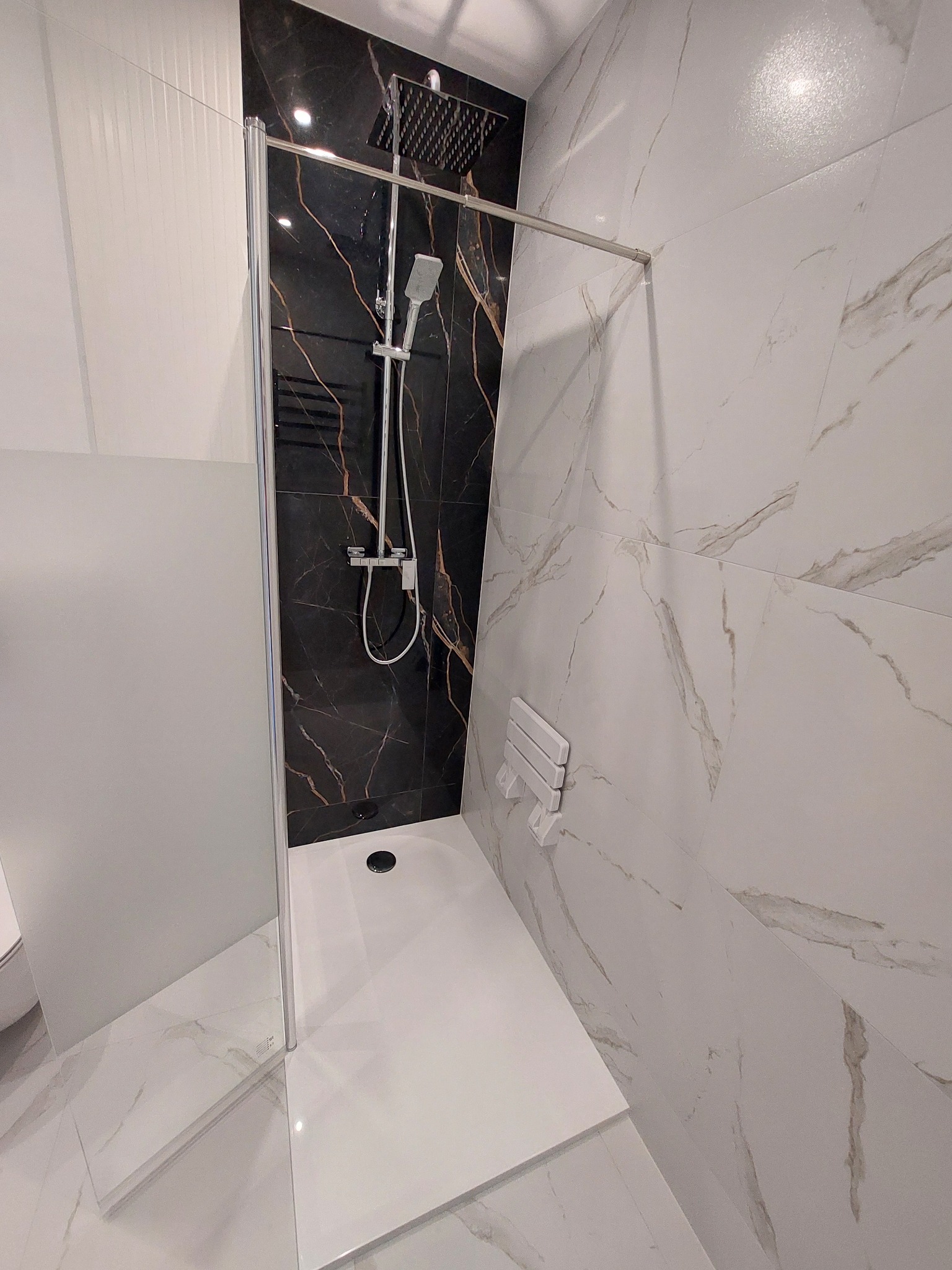 Nowoczesna kabina prysznicowa z czarną ścianą imitującą marmur i białymi płytkami z szarymi żyłami. Minimalistyczny design, chromowana armatura.