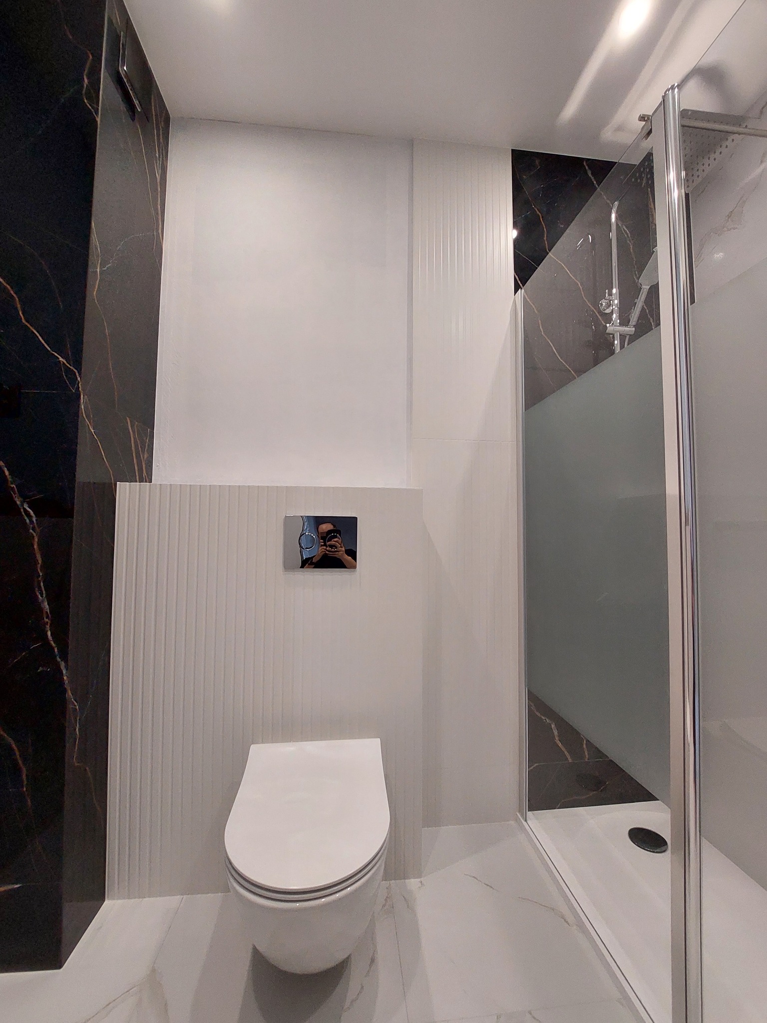 Nowoczesna łazienka z kabiną prysznicową i podwieszaną toaletą. Czarno-białe płytki imitujące marmur. Widoczny fragment odbicia fotografa w przycisku spłuczki.