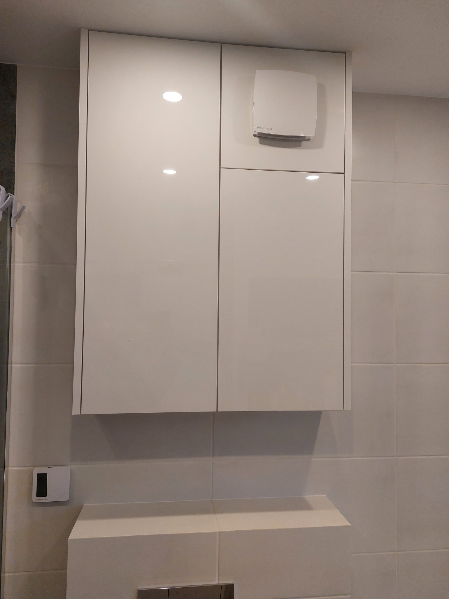 Biała szafka łazienkowa z wbudowanym oświetleniem i wentylatorem na tle jasnych płytek. Nowoczesny design, symetryczny układ.