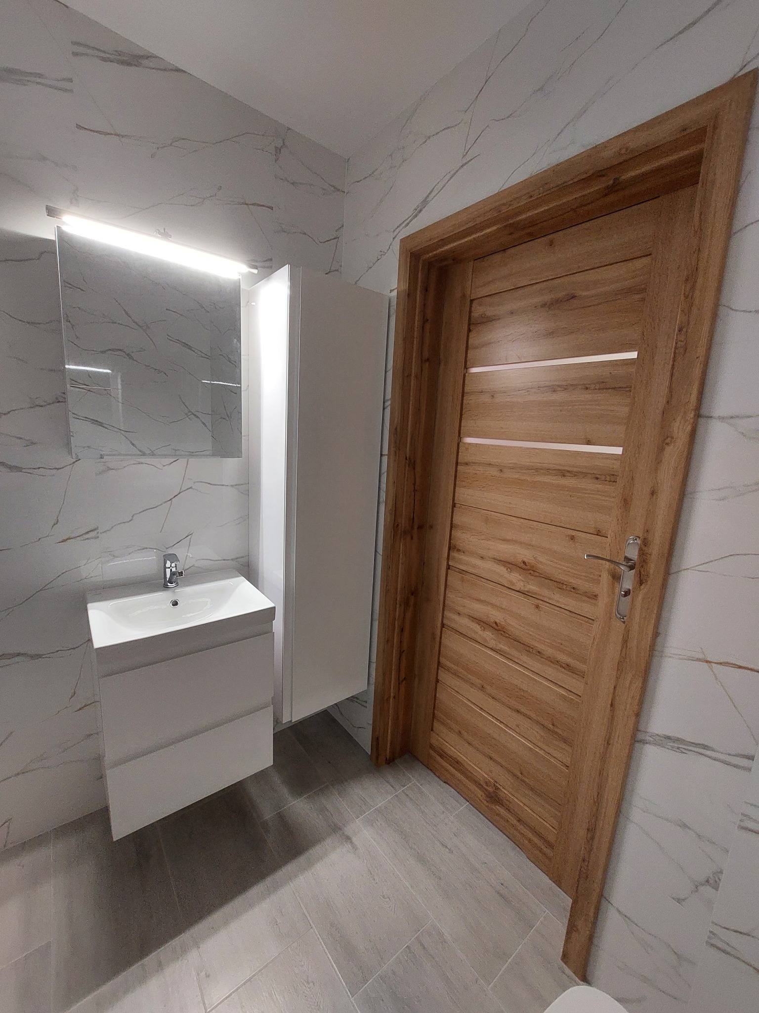 Nowoczesna łazienka po remoncie: biała umywalka z szafką, lustro z oświetleniem LED, drewniane drzwi z poziomymi wstawkami i marmurowe płytki na ścianach i podłodze.