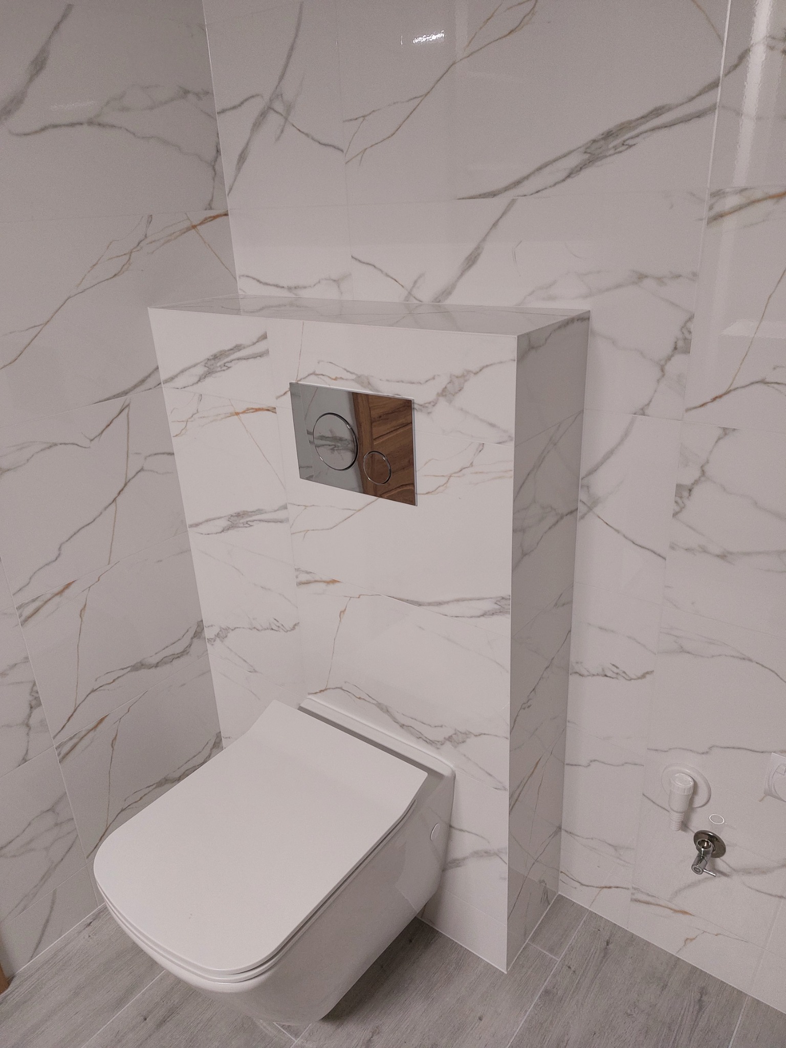 Podwieszana toaleta z zabudowaną spłuczką na tle płytek imitujących marmur z delikatnymi złotymi żyłami. Chromowany przycisk spłukujący.