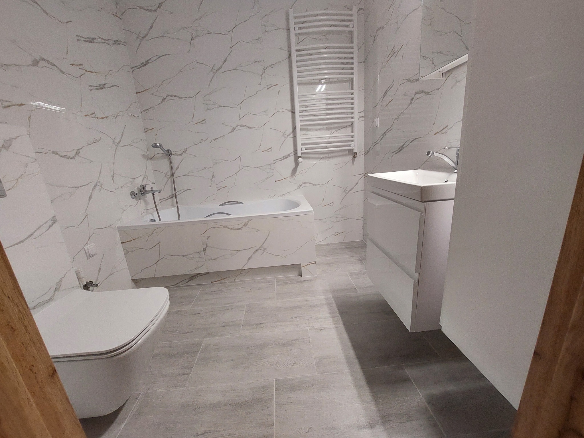 Nowoczesna, wykończona łazienka z płytkami imitującymi marmur, z białą armaturą i grzejnikiem drabinkowym. Podłoga wyłożona płytkami w odcieniach szarości.