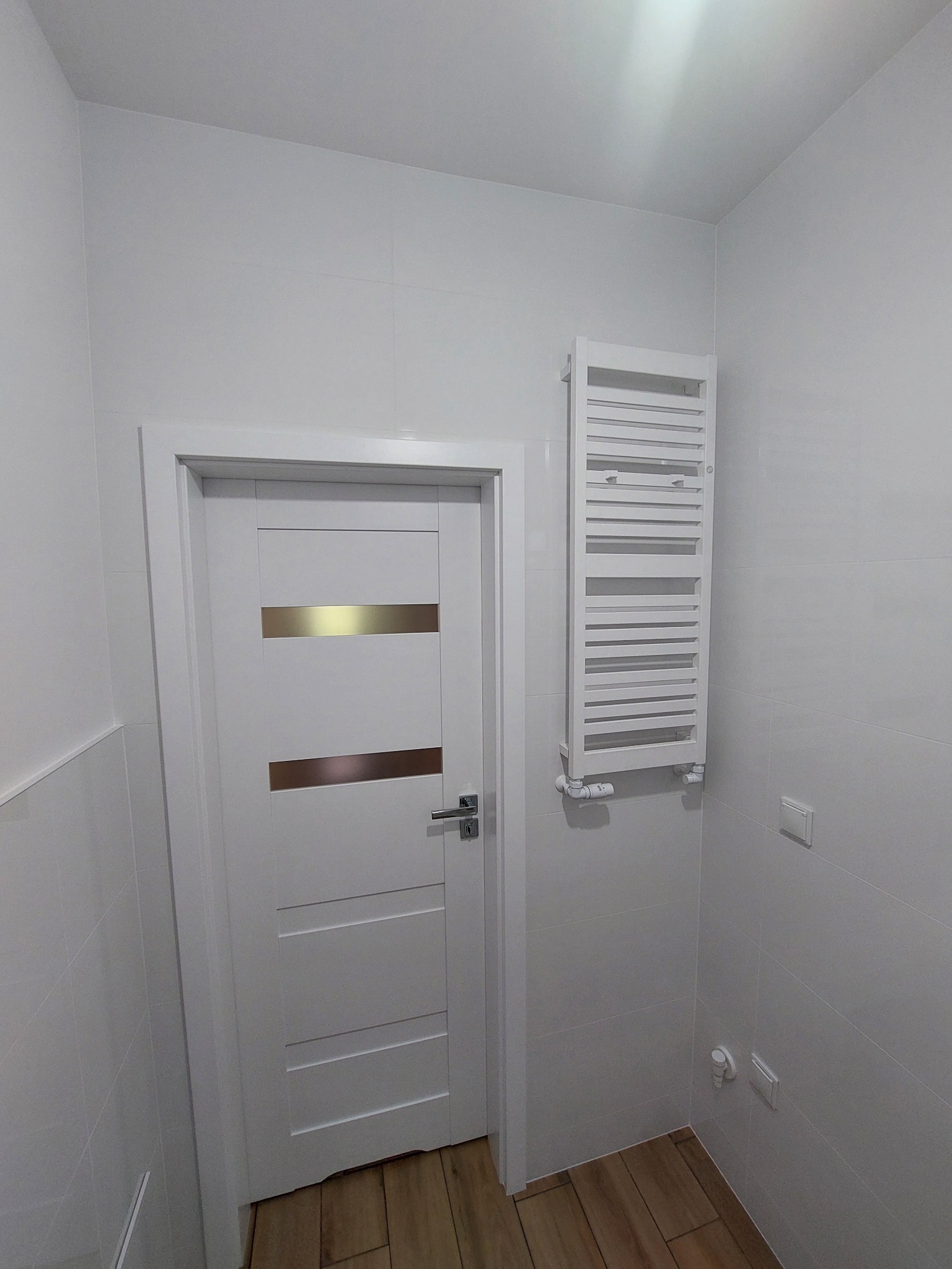 Biała łazienka po remoncie: płytki na ścianach, drewnopodobna podłoga, grzejnik drabinkowy i białe drzwi z poziomymi, złotymi paskami.