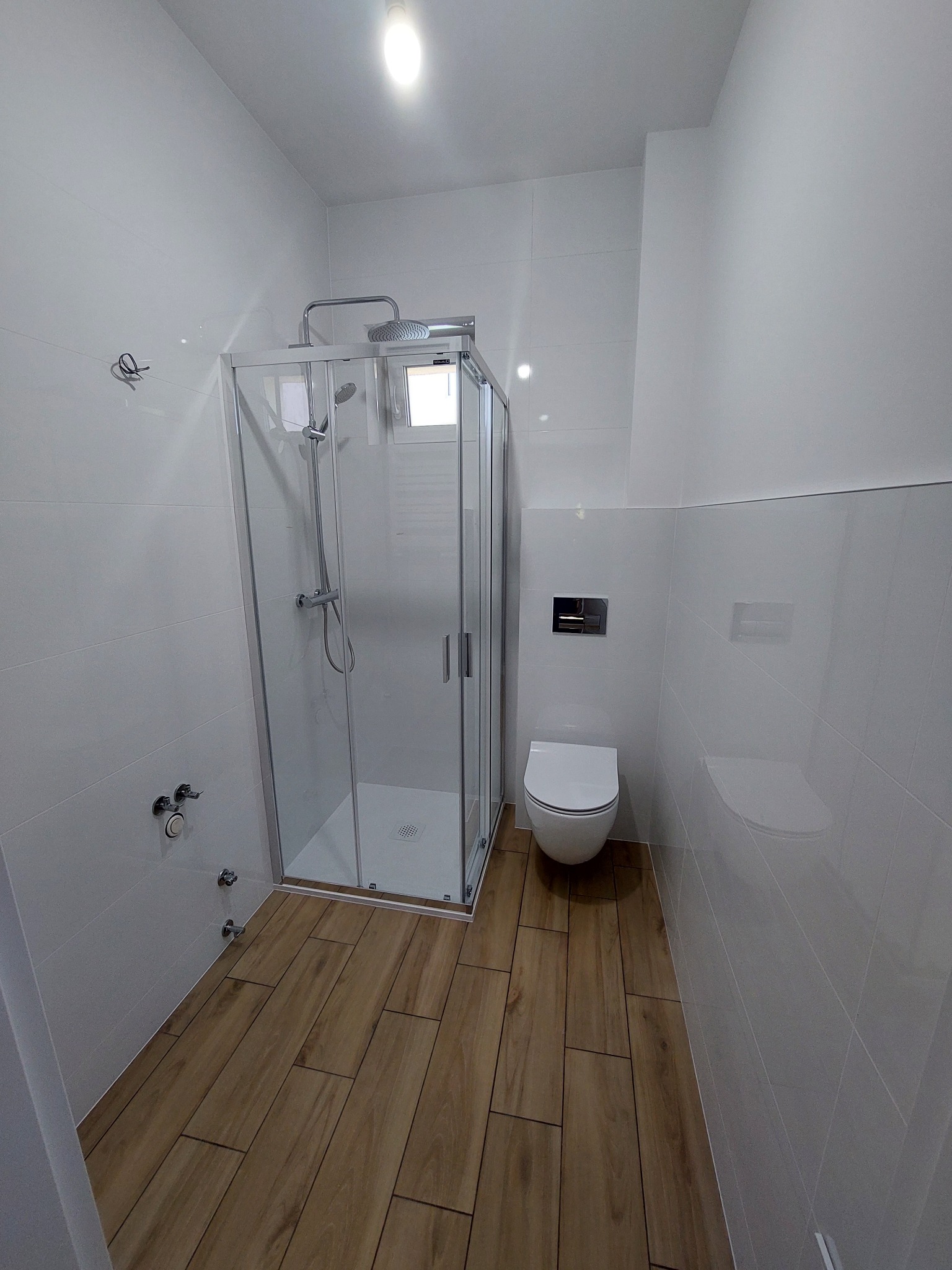 Nowoczesna łazienka z kabiną prysznicową, toaletą podwieszaną i drewnopodobną podłogą. Ściany wyłożone białymi, prostokątnymi płytkami.