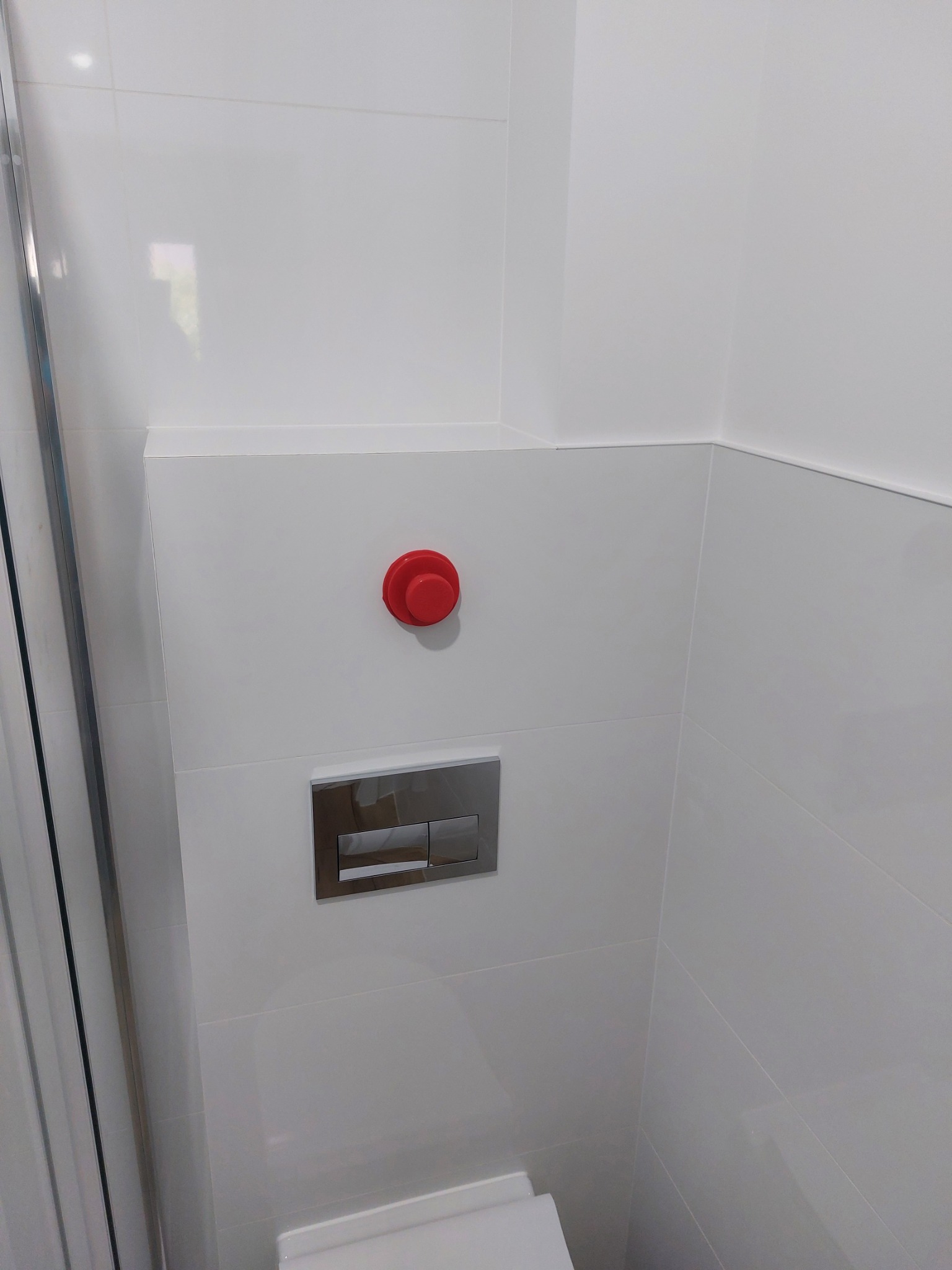 Biała, wykafelkowana ściana w łazience z chromowanym przyciskiem spłukiwania toalety i czerwonym odpowietrznikiem. Widoczny fragment kabiny prysznicowej.