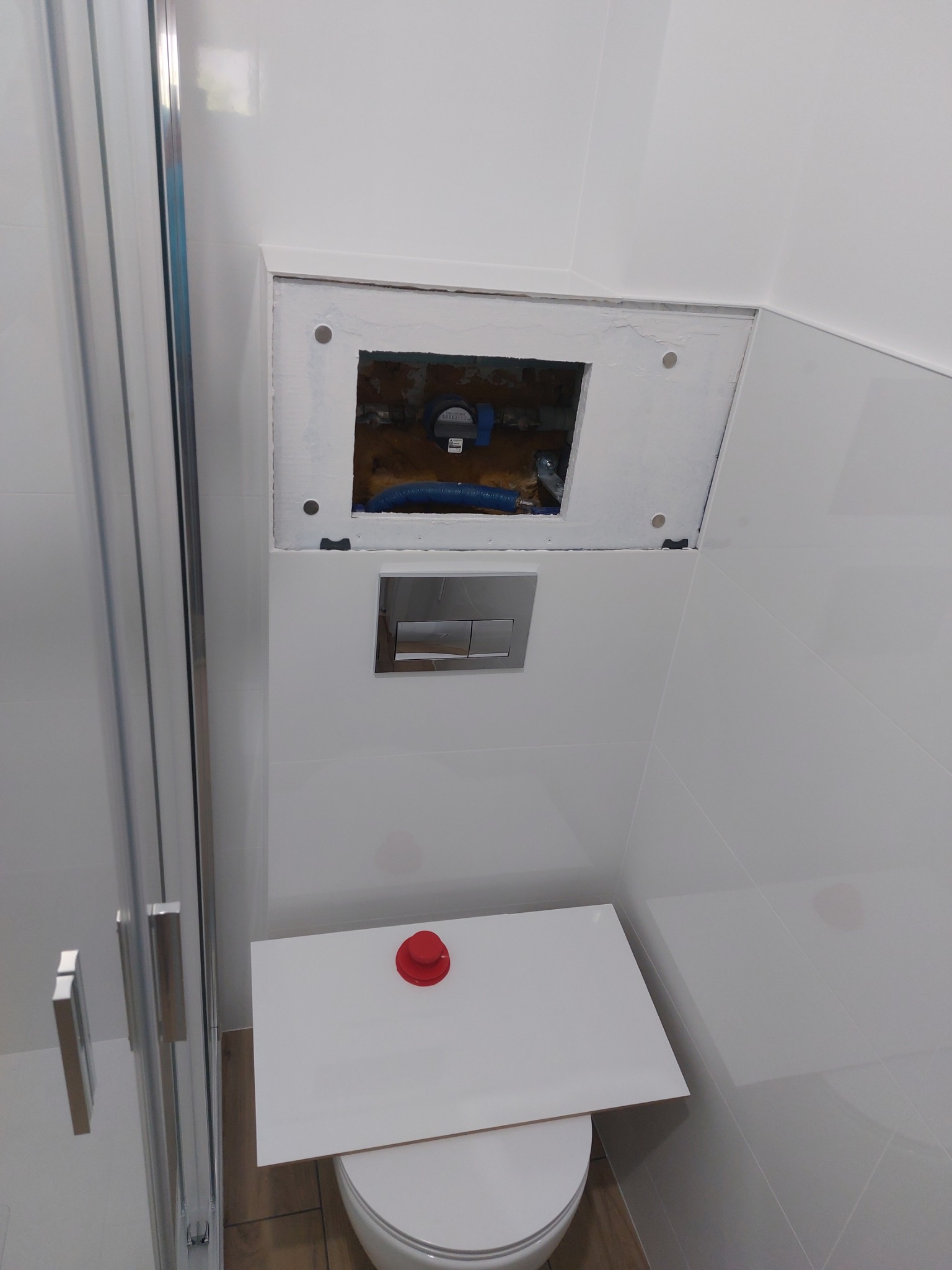 Biała łazienka z widoczną instalacją podtynkową w otworze rewizyjnym, nad spłuczką WC. Widoczny fragment kabiny prysznicowej.