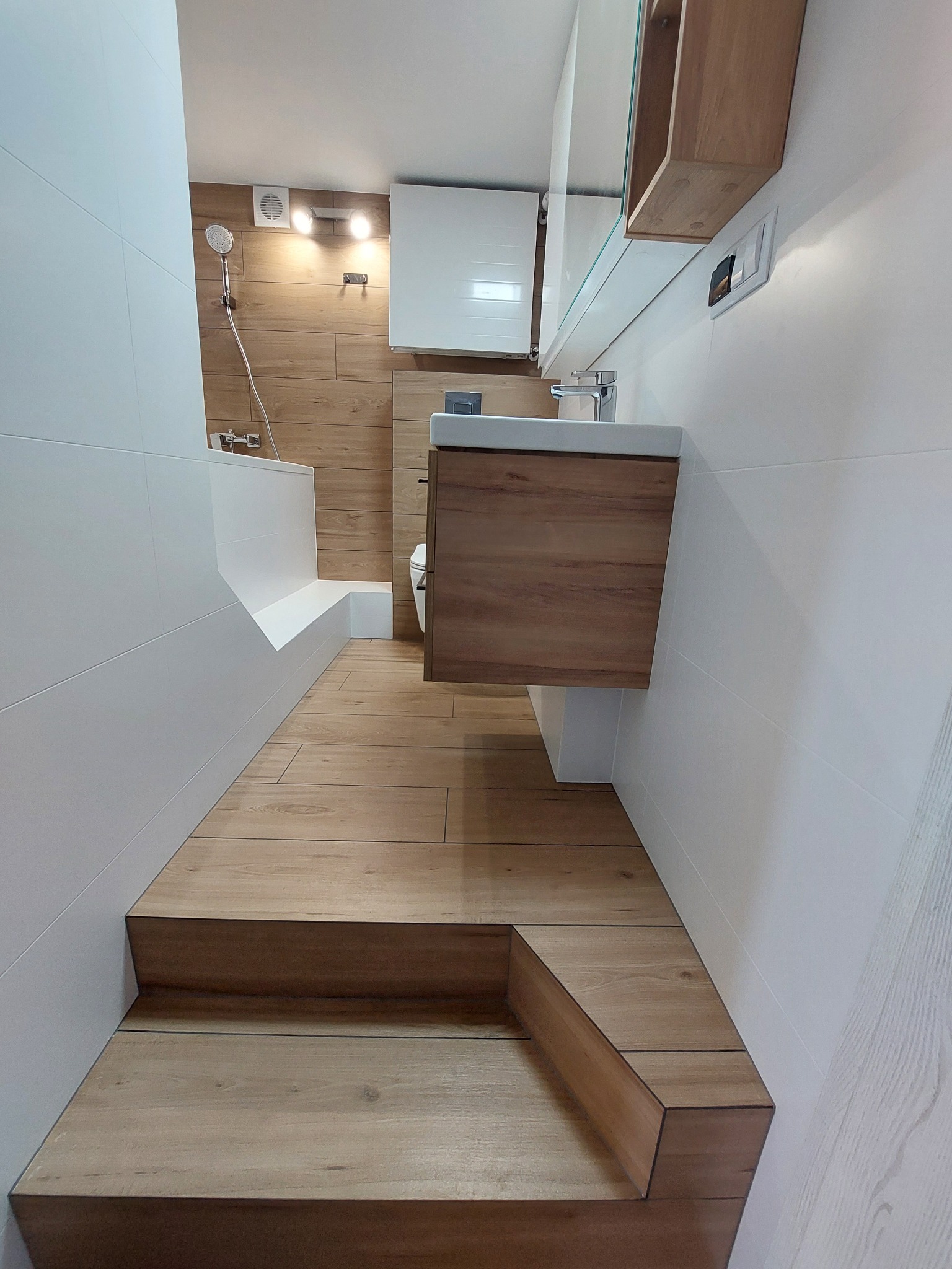 Nowoczesna łazienka z drewnopodobnymi płytkami na podłodze i ścianie prysznicowej, białe płytki na pozostałych ścianach, zabudowana wanna i szafka z umywalką.