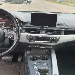 Wnętrze samochodu Audi A4 z widocznym kokpitem, kierownicą z logo Audi, wyświetlaczem, konsolą środkową i elementami wykończenia w kolorze srebrnym.