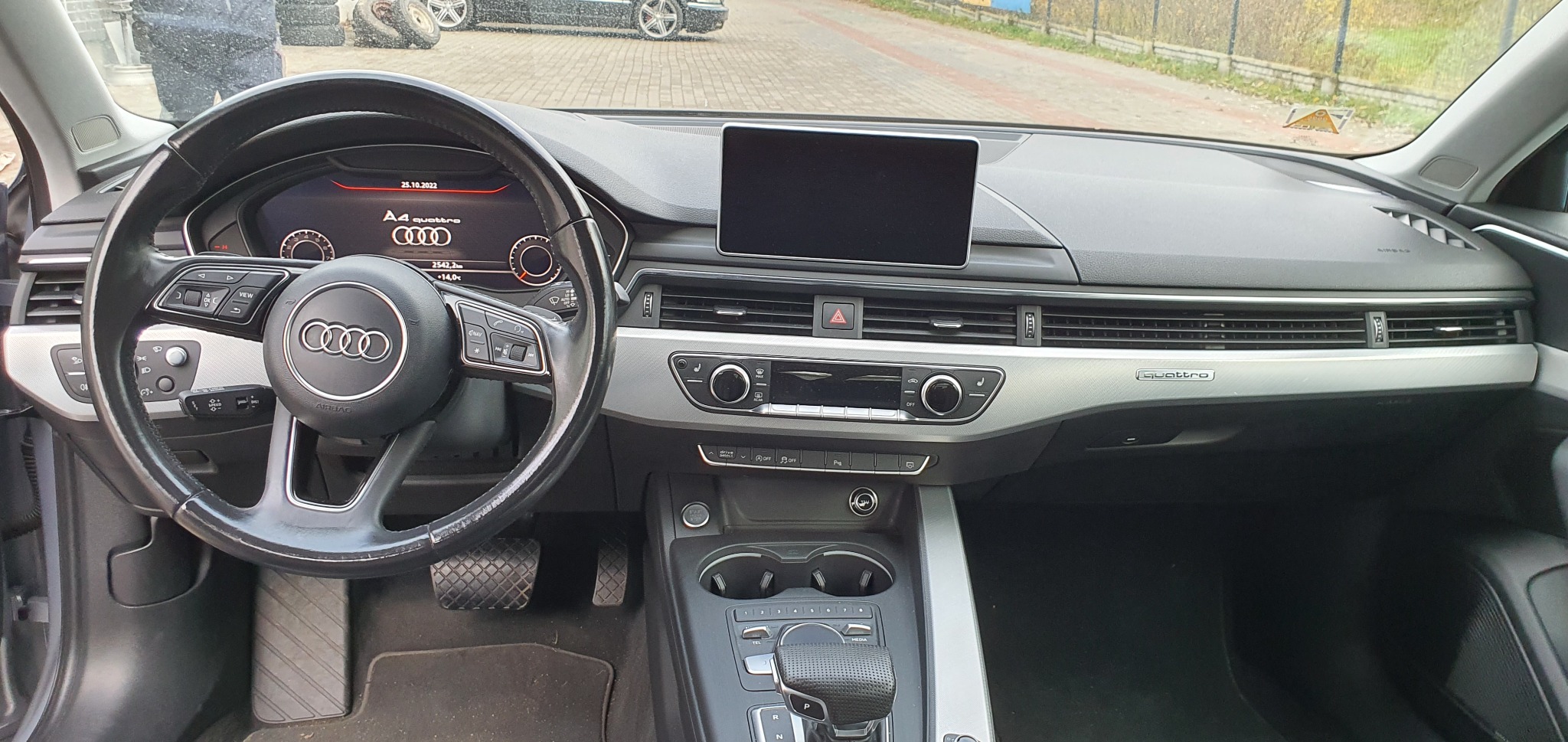 Wnętrze samochodu Audi A4 z widocznym kokpitem, kierownicą z logo Audi, wyświetlaczem, konsolą środkową i elementami wykończenia w kolorze srebrnym.