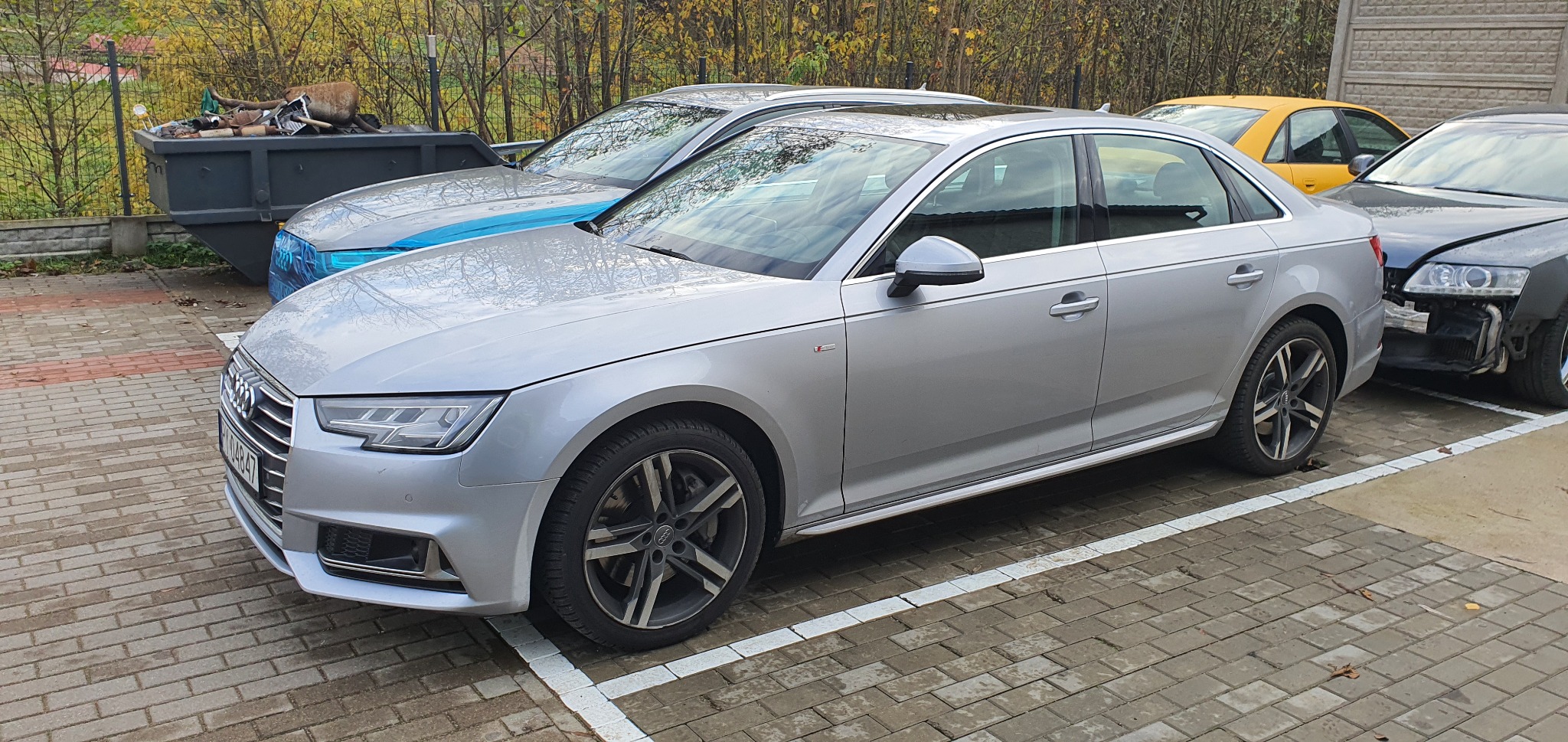 Srebrny sedan Audi A4 zaparkowany na brukowanym parkingu, obok uszkodzonego samochodu z widocznymi elementami zawieszenia i żółtego auta w tle, z kontenerem na śmieci w oddali.