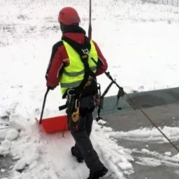 Pracownik w kasku i kamizelce odblaskowej, zabezpieczony linami, odśnieża płaski dach za pomocą czerwonej łopaty na kółkach; w tle ośnieżone pole i budynek przemysłowy.