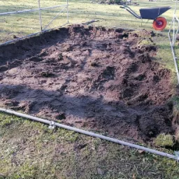 Wykopana prostokątna dziura w ziemi, otoczona metalową ramą, z taczką w tle.