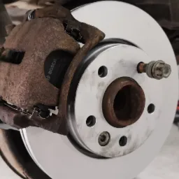 Wymiana tarczy hamulcowej: nowa, srebrna tarcza założona, widoczny skorodowany zacisk hamulcowy z klockami Bosch, auto na podnośniku.