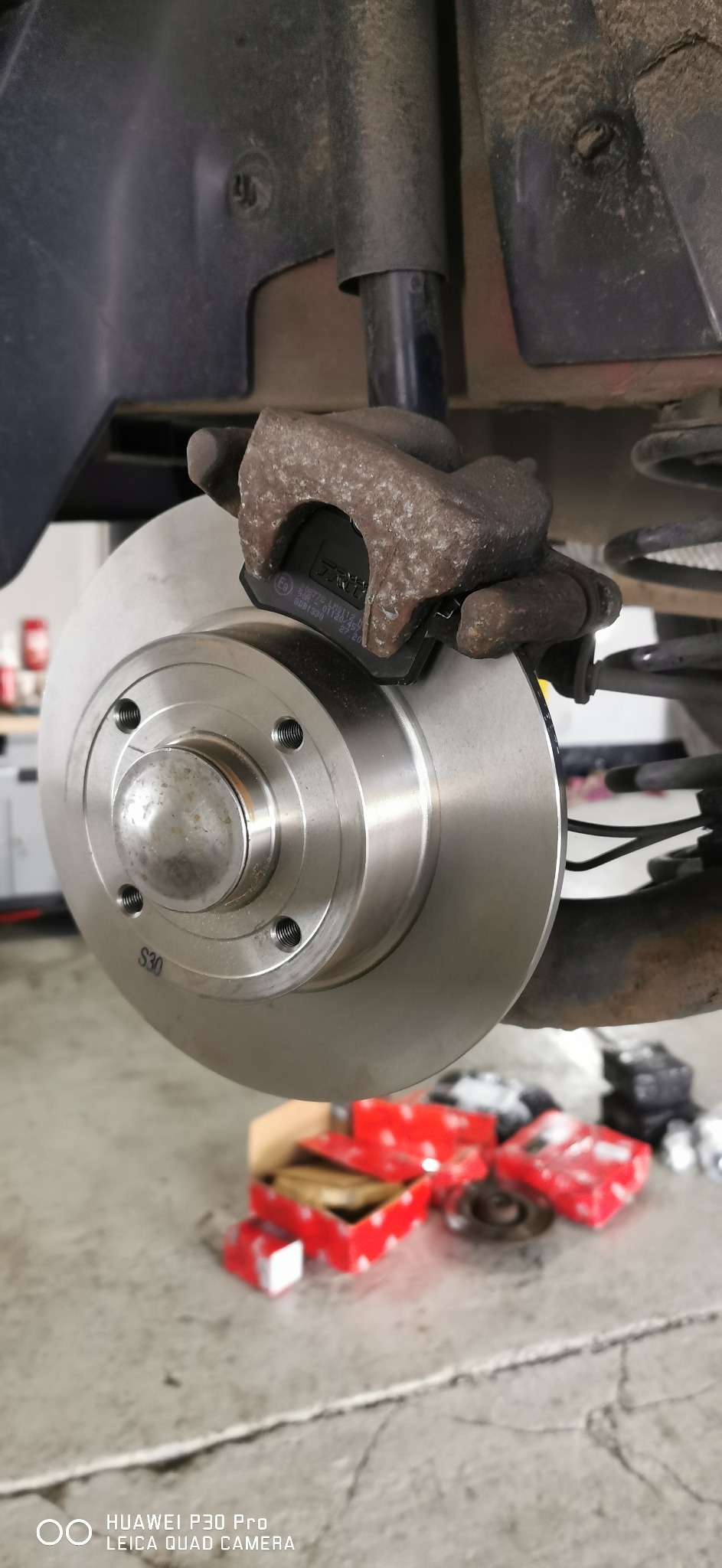 Nowa tarcza hamulcowa z zaciskiem i klockami, widoczne pudełka po częściach zamiennych na podłodze warsztatu.