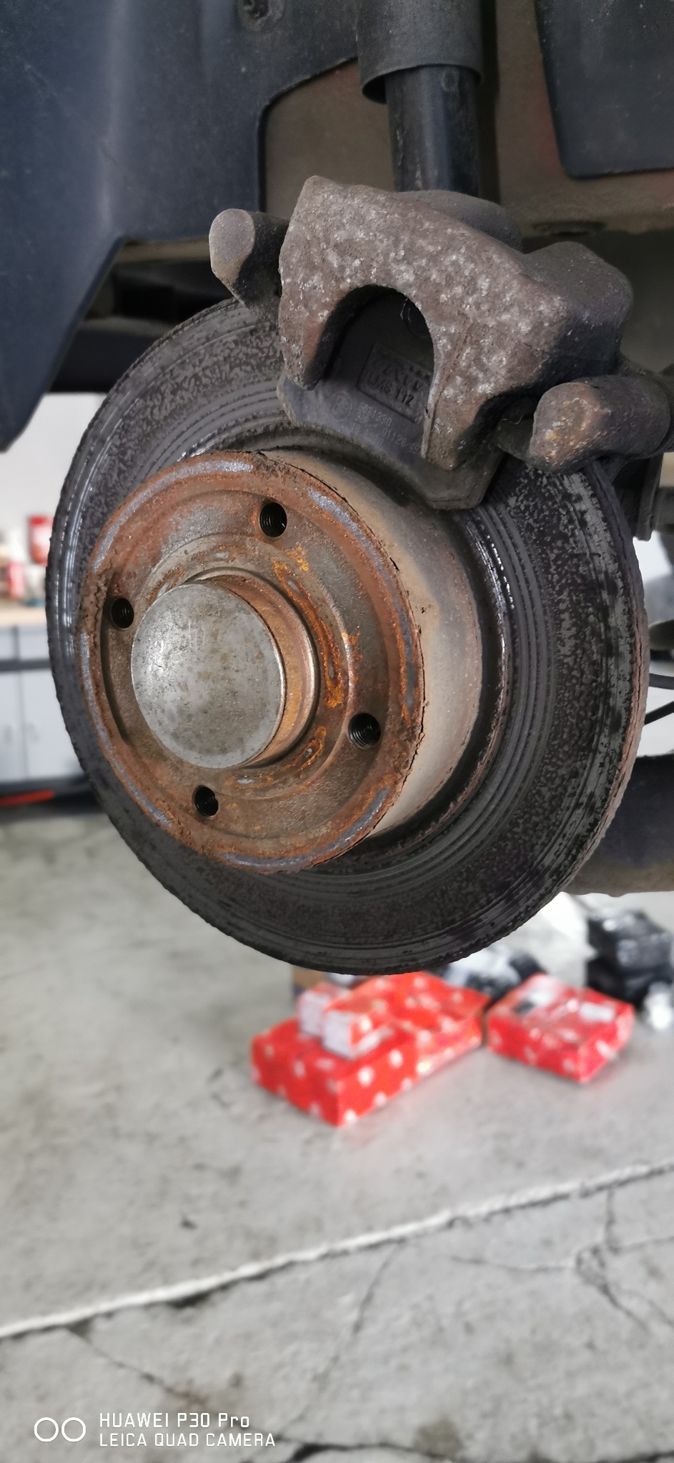 Zużyta tarcza hamulcowa z widoczną korozją i zaciskiem hamulcowym, w tle pudełka z nowymi częściami zamiennymi.