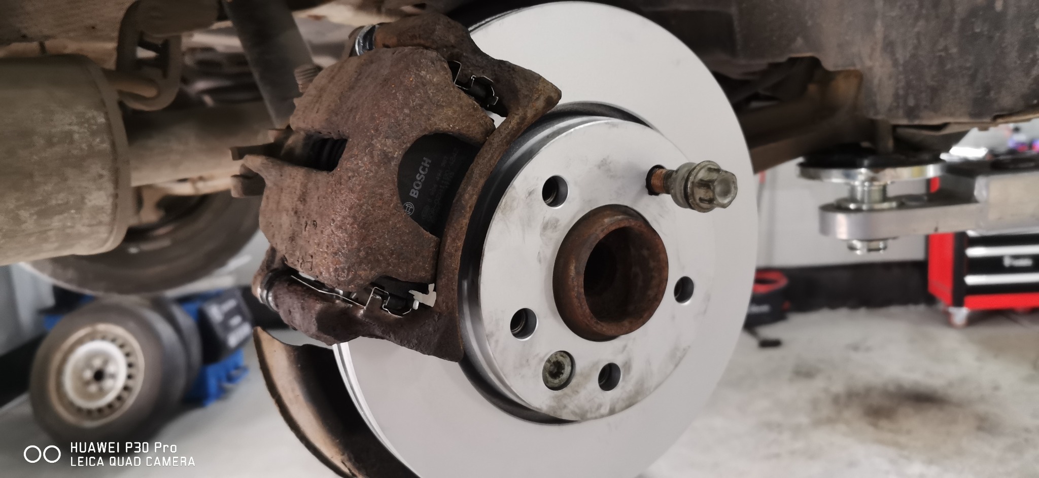 Wymiana tarczy hamulcowej: nowa, srebrna tarcza założona, widoczny skorodowany zacisk hamulcowy z klockami Bosch, auto na podnośniku.