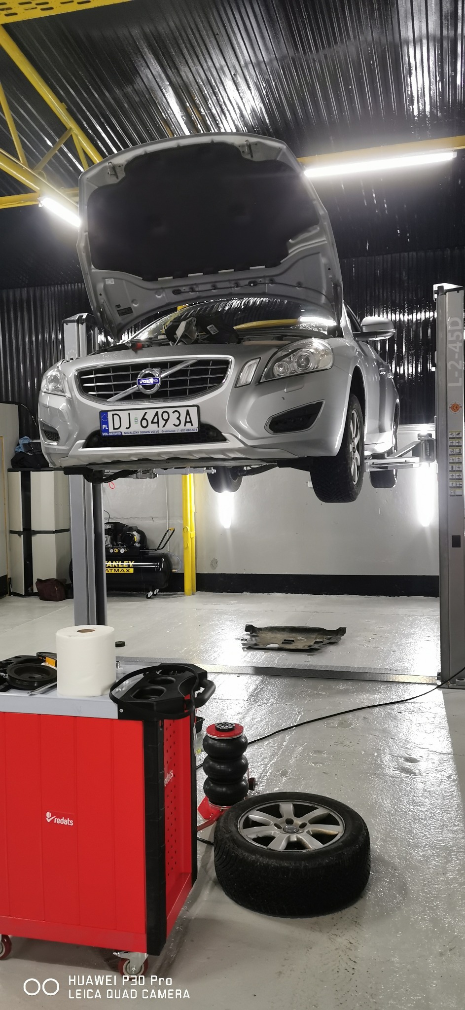 Srebrny samochód marki Volvo z otwartą maską, podniesiony na podnośniku w warsztacie samochodowym, widoczny numer rejestracyjny, obok leży zdjęte koło, w tle kompresor i narzędzia.