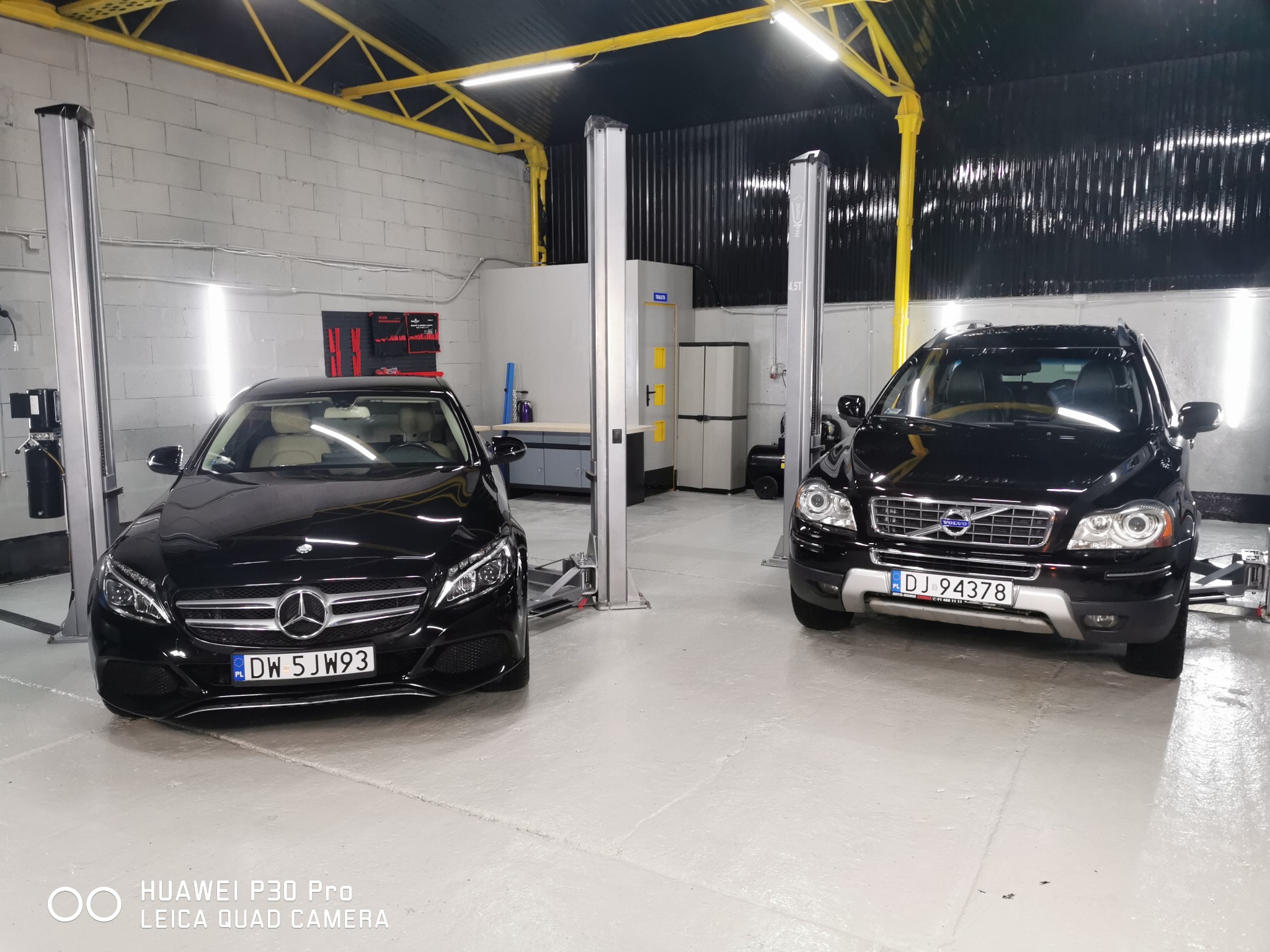 Dwa czarne samochody (Mercedes i Volvo) w warsztacie, każdy podnośniku samochodowym; widoczne tablice rejestracyjne.