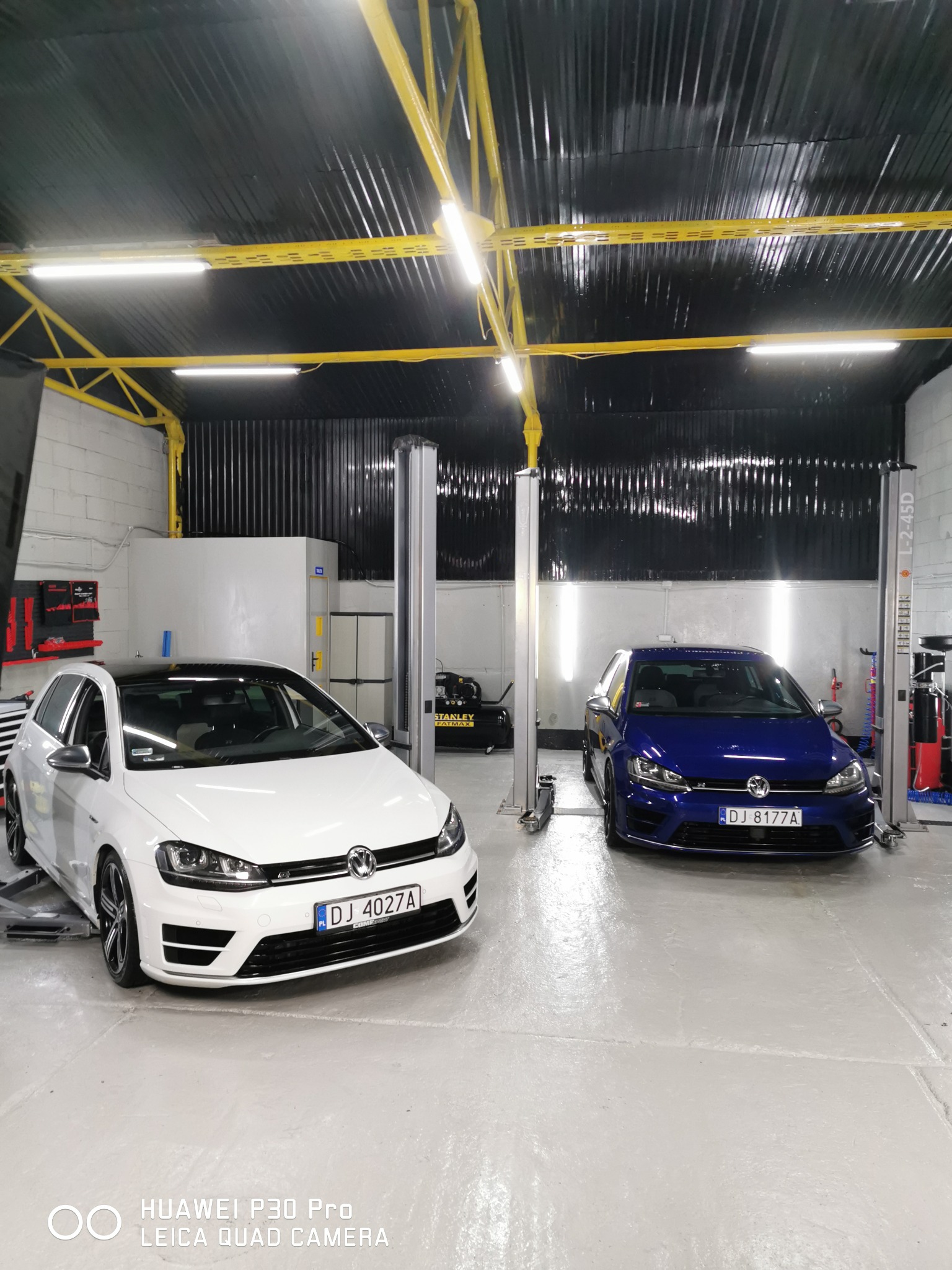 Wnętrze warsztatu samochodowego z dwoma samochodami Volkswagen Golf R na podnośnikach: biały z czarnym dachem po lewej i niebieski po prawej, pod żółtą konstrukcją sufitu i lampami.
