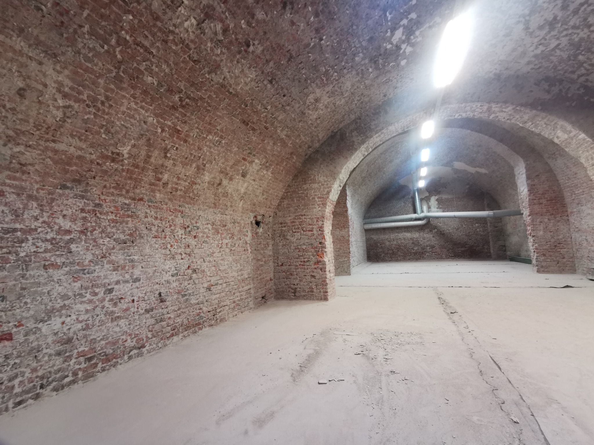 Ceglane sklepienie piwniczne z widocznymi śladami renowacji, surowy stan wykończenia, posadzka pokryta warstwą pyłu, oświetlenie jarzeniowe.