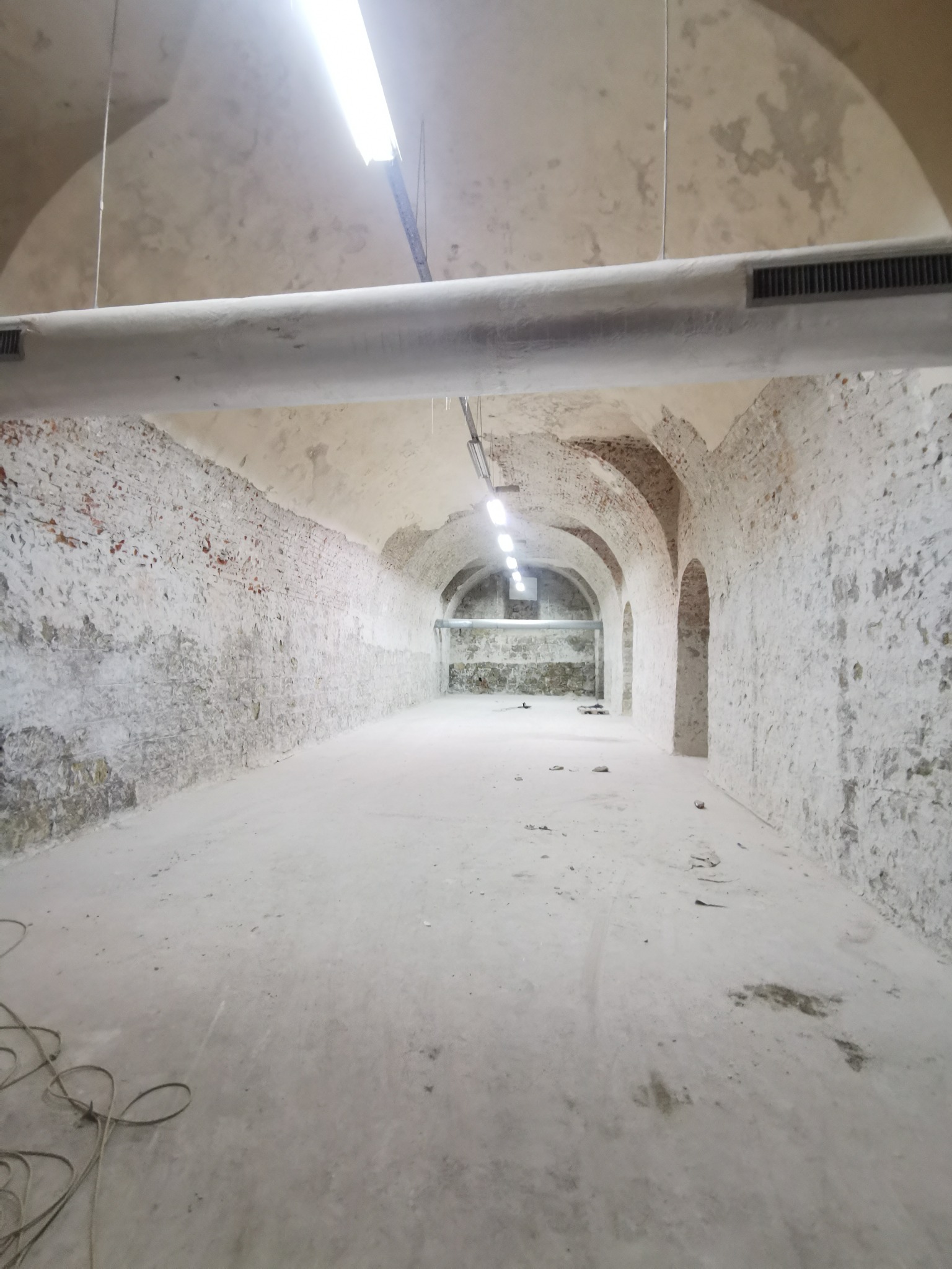 Wnętrze piwnicy z łukowatym sufitem i ścianami z cegły pokrytymi białym nalotem, oświetlone jarzeniówkami, z widocznymi śladami po wcześniejszych pracach.